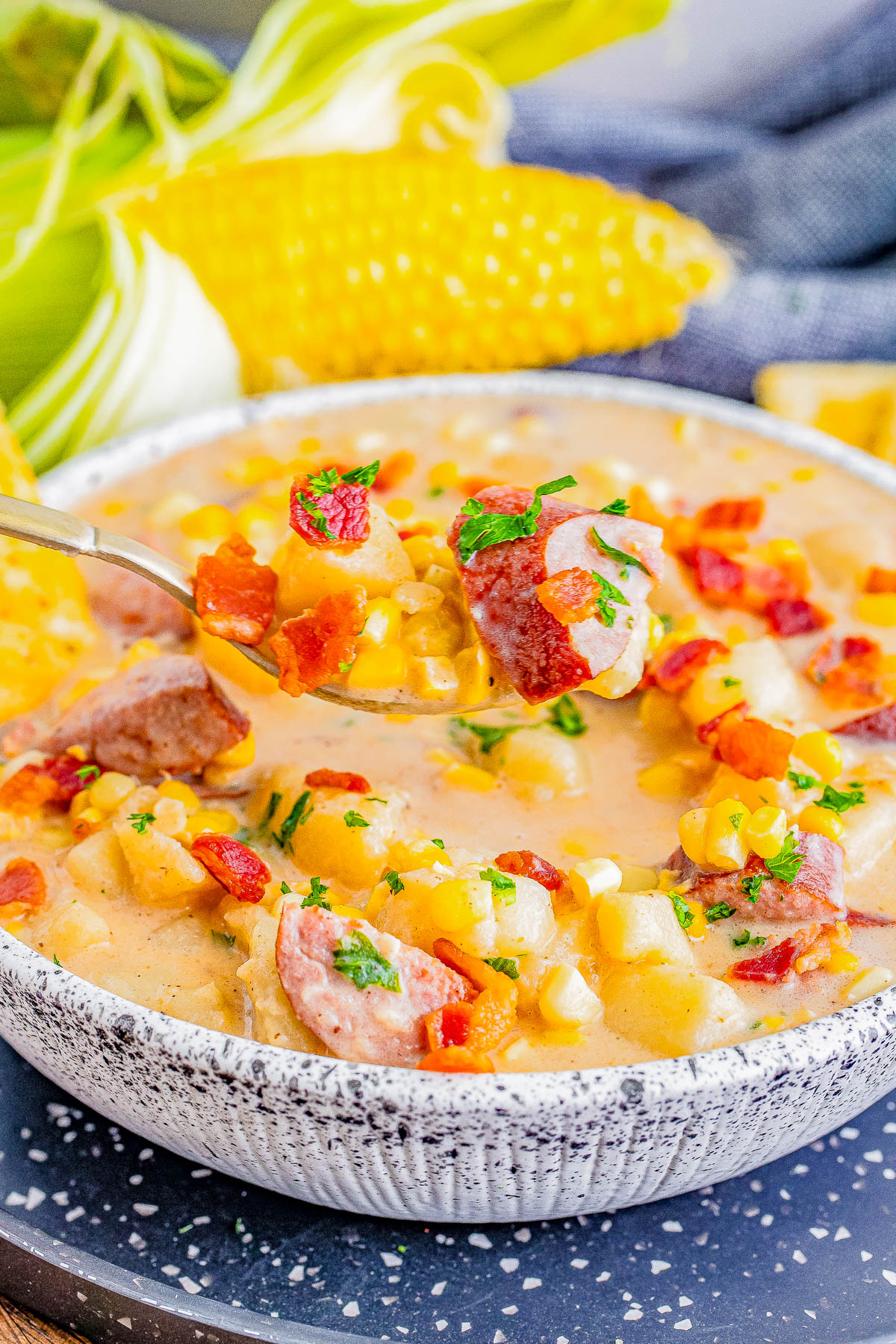 A spoonful of creamy corn chowder with sausage and bacon pieces is lifted from a bowl, garnished with herbs. Corn on the cob can be seen in the background.