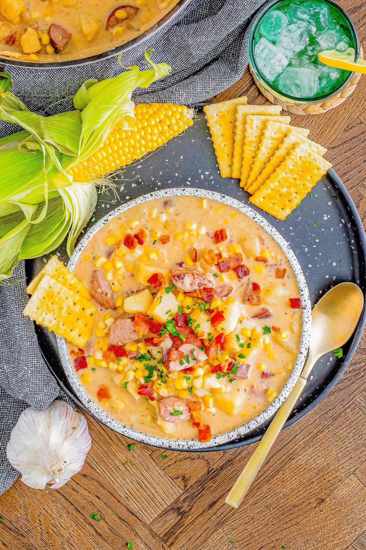 A bowl of corn chowder topped with bacon and herbs is served with crackers and a corn cob on a black plate. A garlic bulb and a green drink are beside the plate.