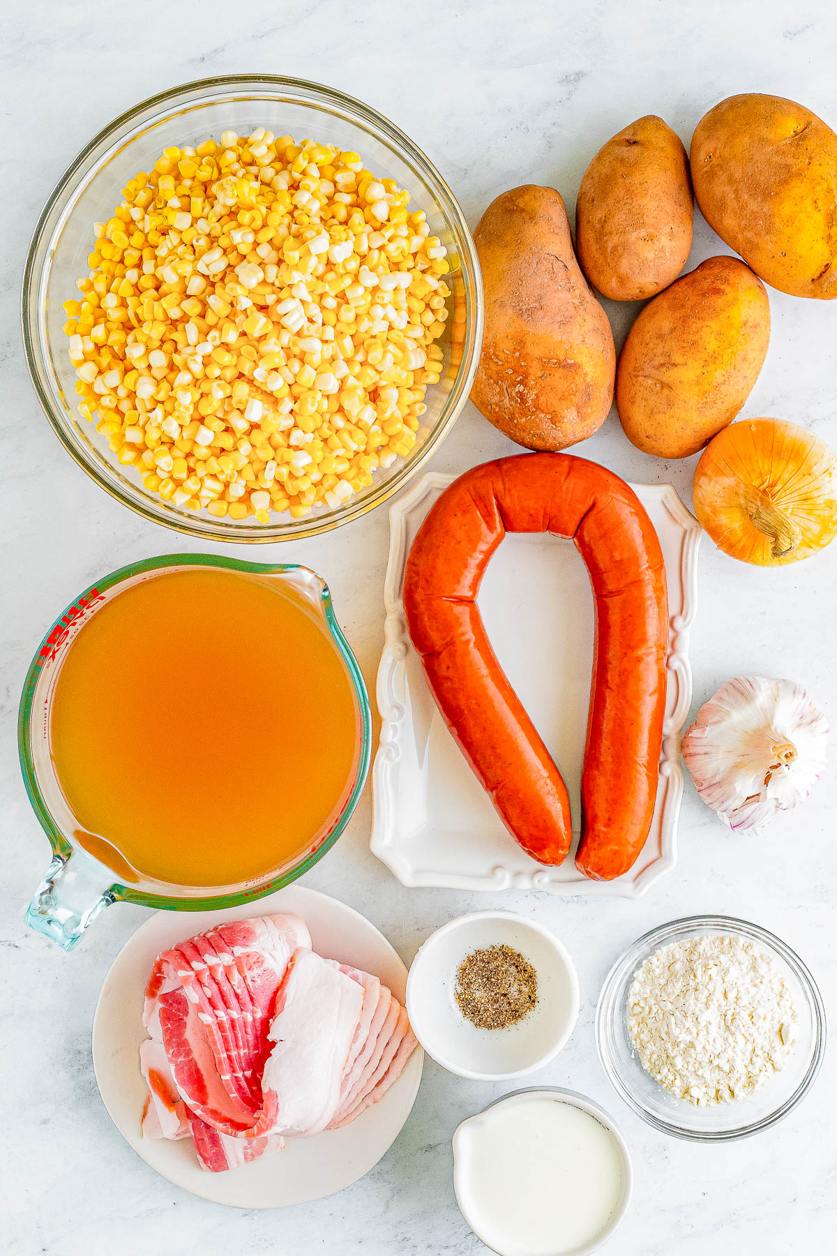 Top-down view of ingredients on a countertop: a bowl of corn, three potatoes, an onion, garlic, sausage, broth in a measuring cup, slices of bacon, a bowl of spices, a bowl of flour, and a small cup of cream.