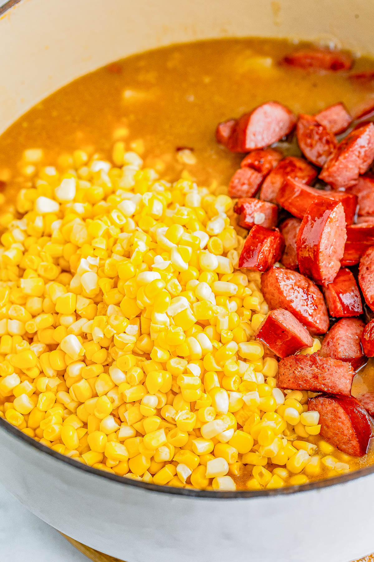 A pot of soup ingredients with corn kernels and sliced sausages in broth.