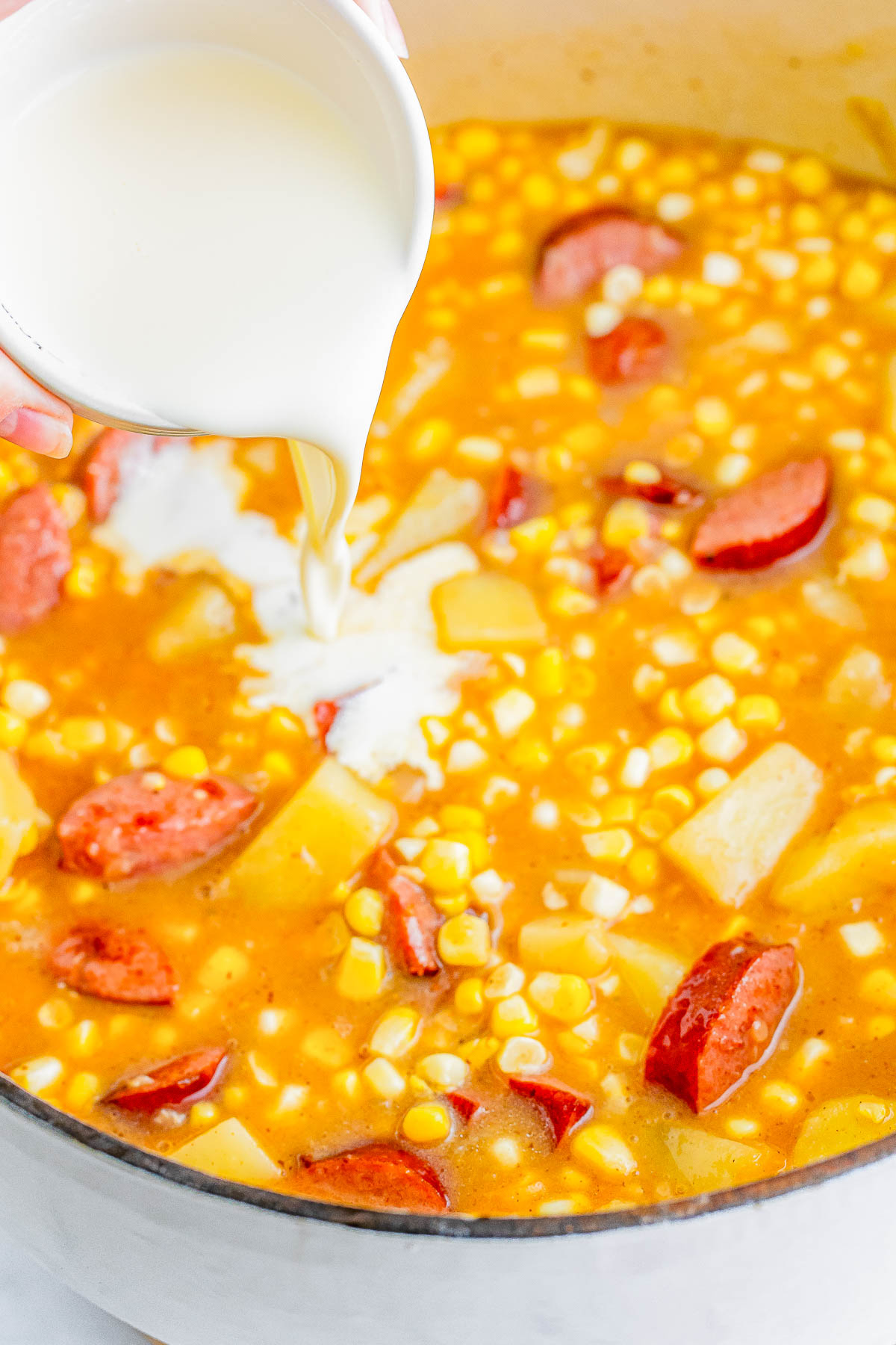 Milk being poured into a pot containing a mixture of corn, potatoes, and sausage in a rich broth.