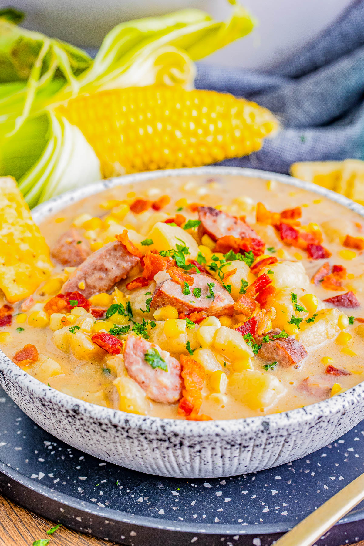 A bowl of creamy corn chowder garnished with bacon pieces and fresh parsley, served with a side of sweet corn and cornbread.