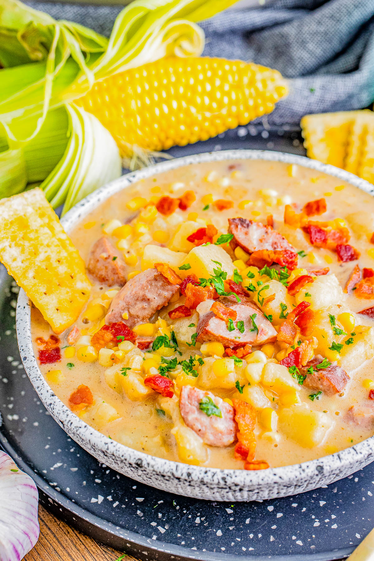 A bowl of corn chowder with pieces of sausage and bacon, garnished with parsley. Fresh corn on the cob and a gray cloth are in the background.