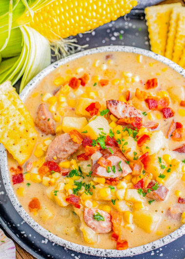 A bowl of creamy corn chowder with sausage pieces and diced potatoes, garnished with chopped parsley and crumbled bacon. Corn on the cob and saltine crackers are placed beside the bowl.