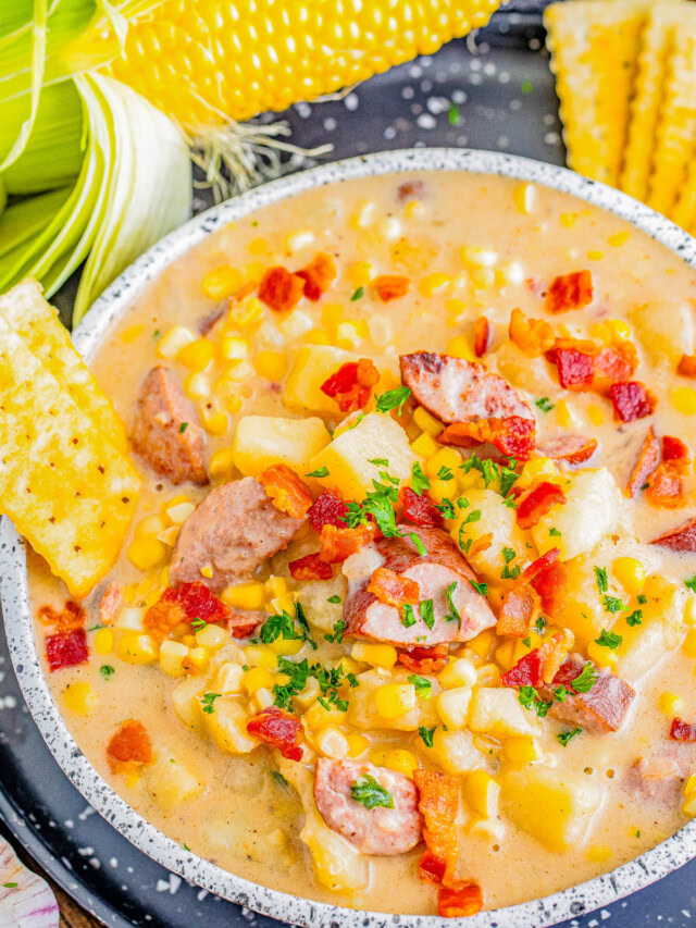 A bowl of creamy corn chowder with sausage pieces and diced potatoes, garnished with chopped parsley and crumbled bacon. Corn on the cob and saltine crackers are placed beside the bowl.