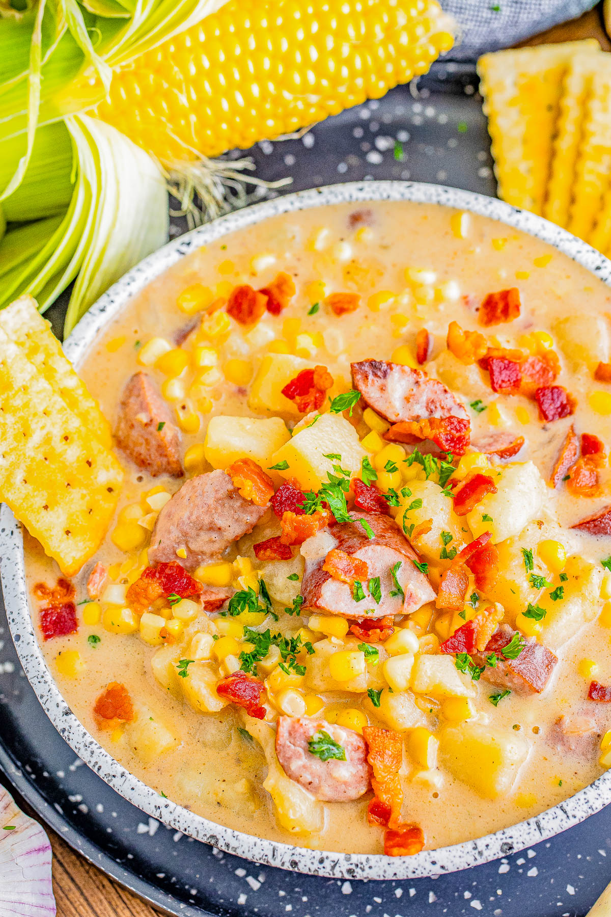 A bowl of creamy corn chowder with sausage pieces and diced potatoes, garnished with chopped parsley and crumbled bacon. Corn on the cob and saltine crackers are placed beside the bowl.