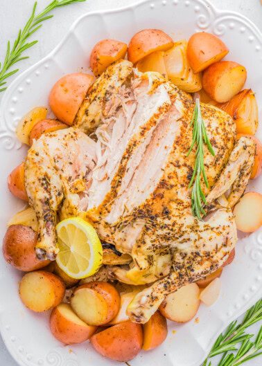 A roasted chicken on a white plate, partially sliced, surrounded by roasted potatoes and garnished with a lemon slice and sprigs of rosemary.