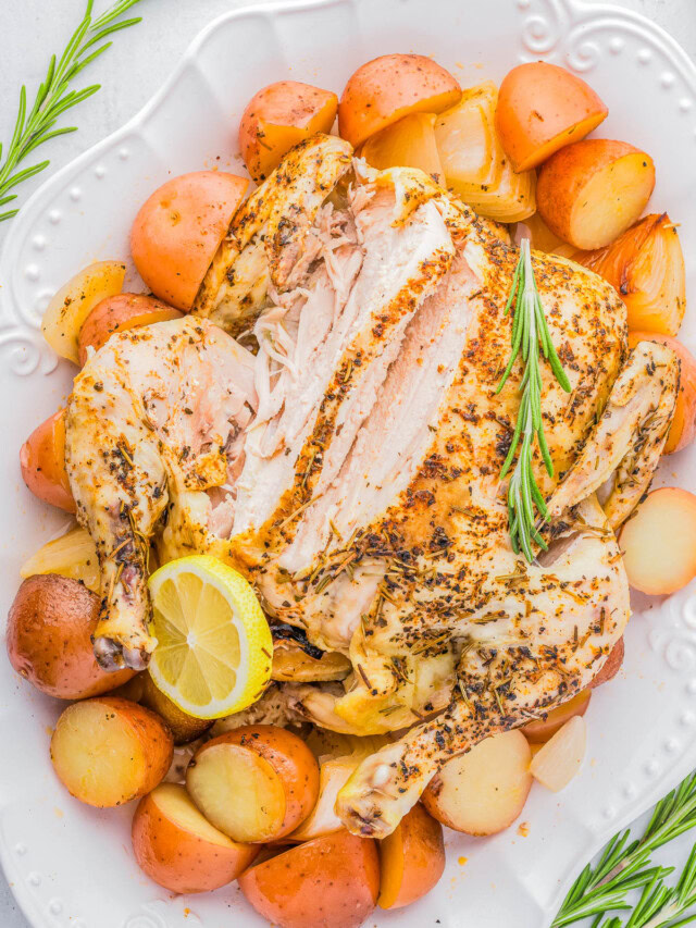 A roasted chicken on a white plate, partially sliced, surrounded by roasted potatoes and garnished with a lemon slice and sprigs of rosemary.