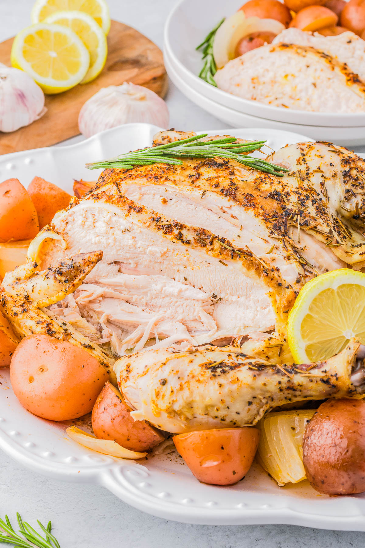 A roasted whole chicken garnished with lemon and rosemary, surrounded by roasted red potatoes and onions on a white plate. In the background are sliced lemons and garlic on a wooden board.