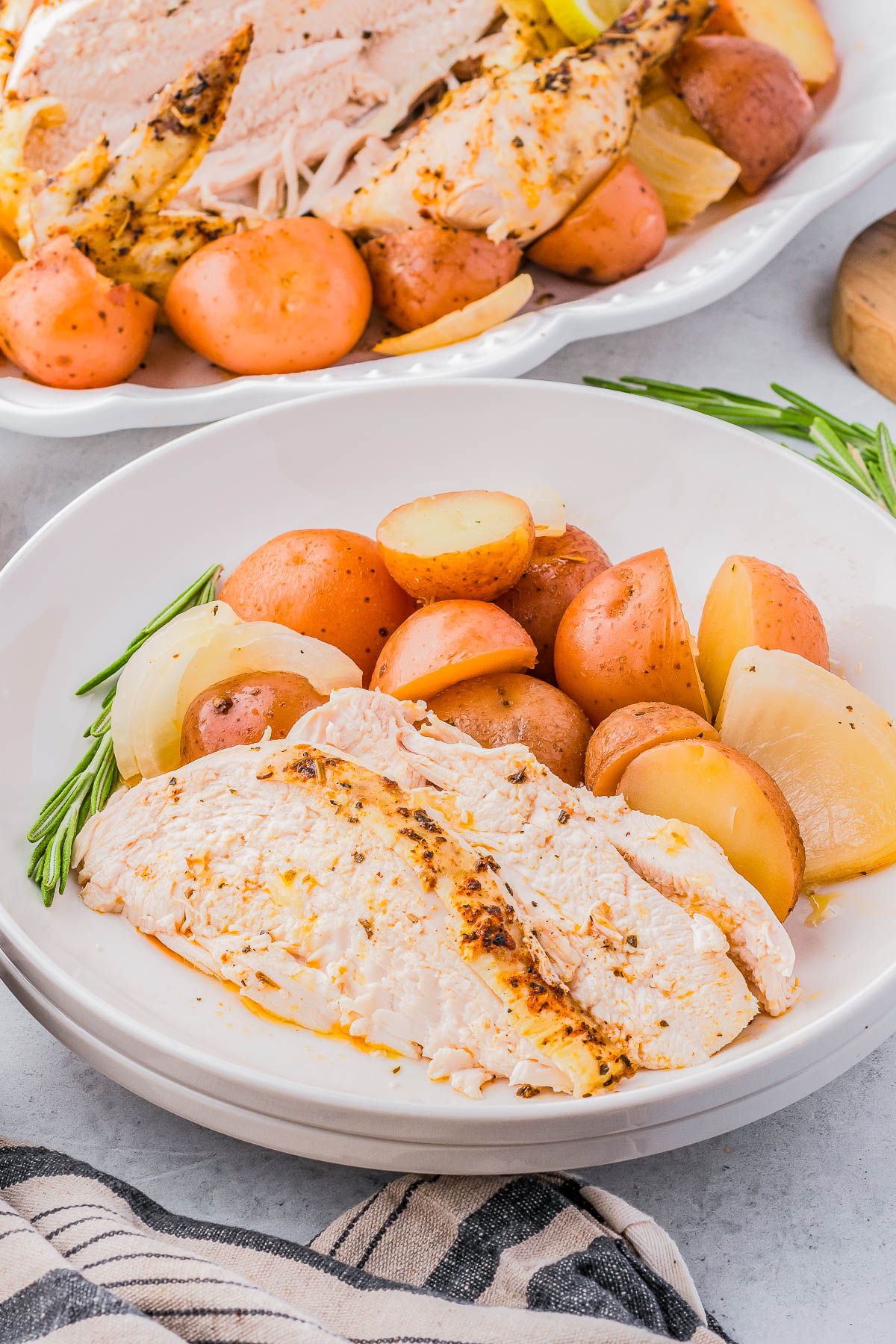 A white plate with slices of roasted chicken breast, red potatoes, and onion, garnished with rosemary. A serving tray with additional roasted chicken and vegetables is visible in the background.