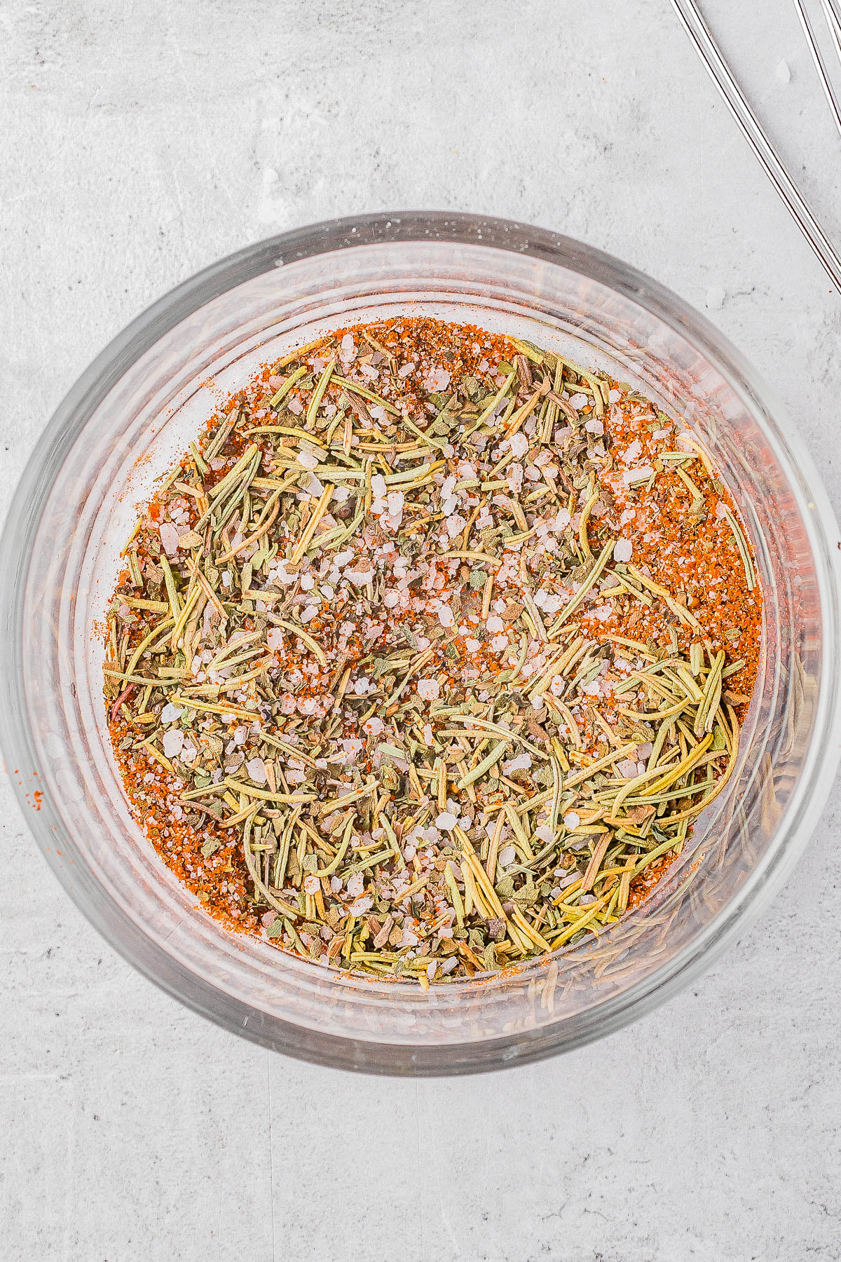 A glass bowl containing a dry spice mixture of rosemary, coarse salt, and orange-brown seasoning on a light gray textured surface.