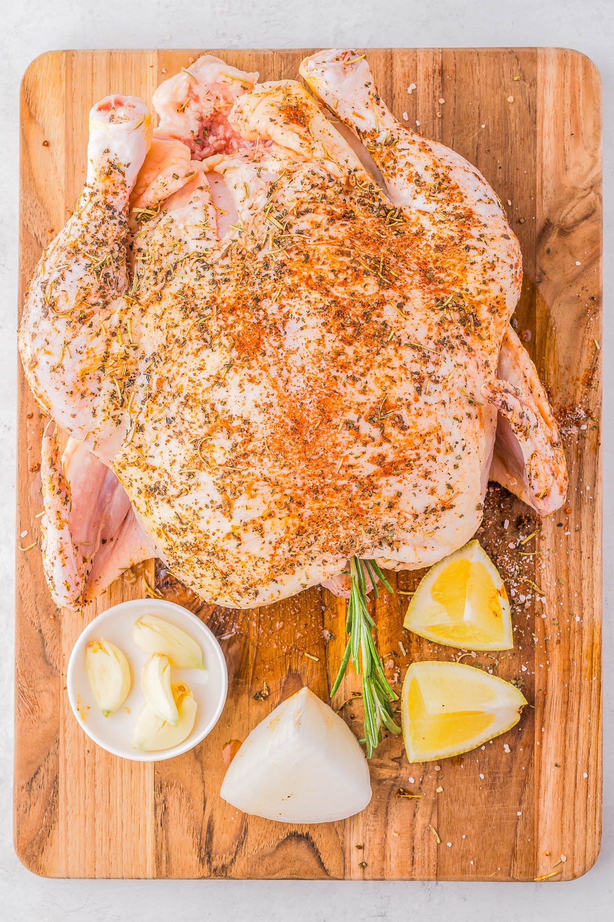 A seasoned whole raw chicken on a wooden cutting board, accompanied by lemon wedges, garlic cloves, and a halved white onion.