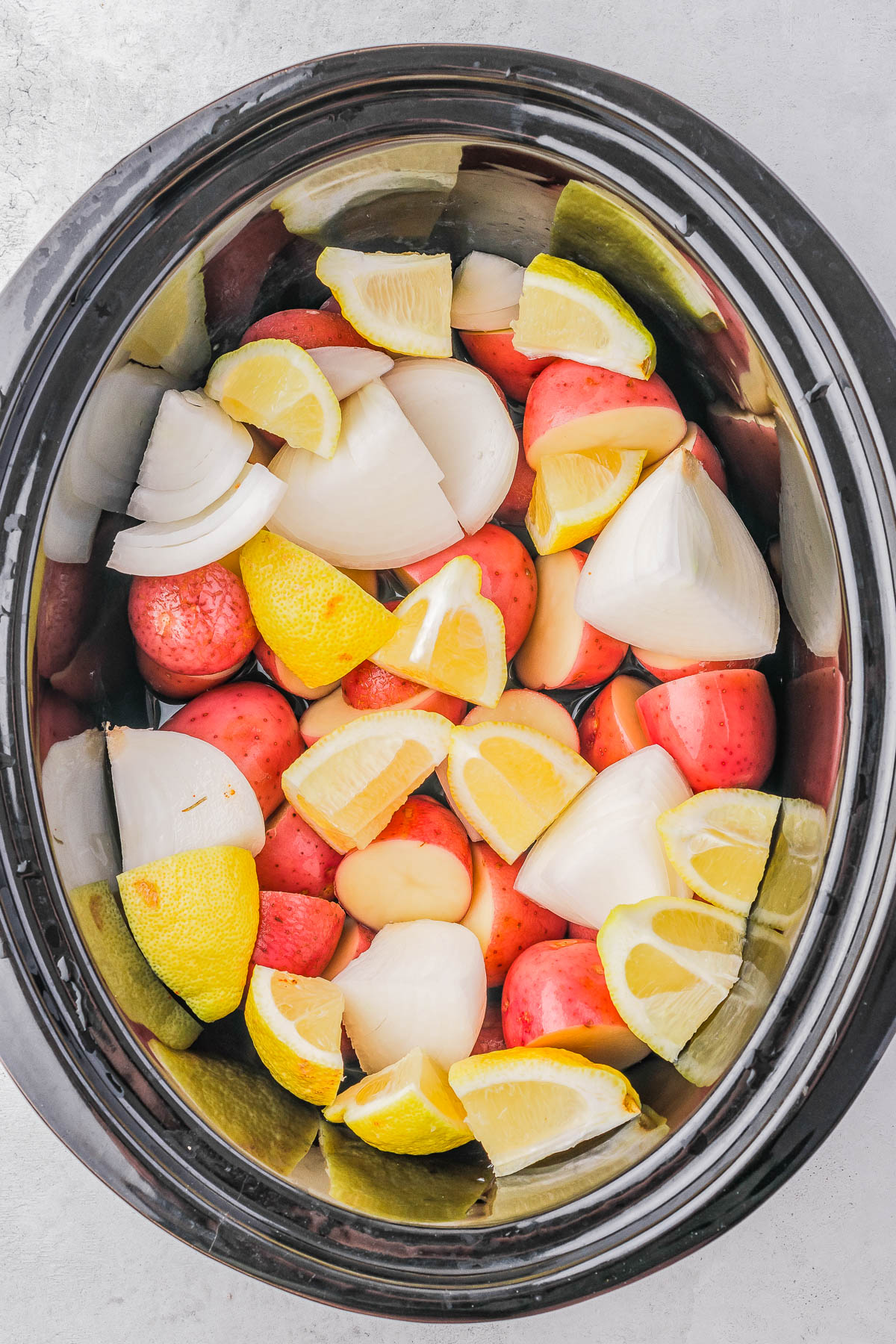 A slow cooker filled with chopped red potatoes, onion wedges, and lemon slices.