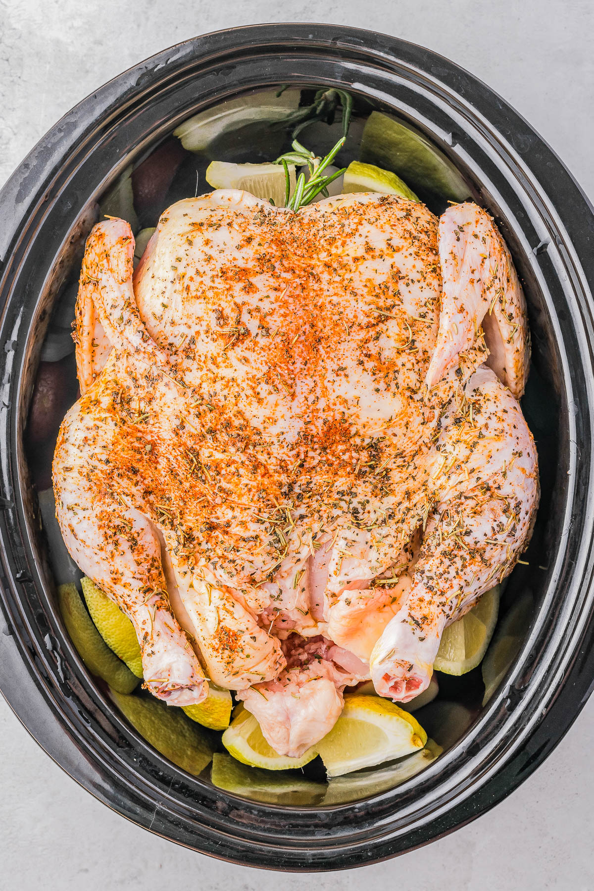 A whole raw chicken seasoned with spices and herbs is placed on top of lemon wedges in a black slow cooker, ready for cooking.