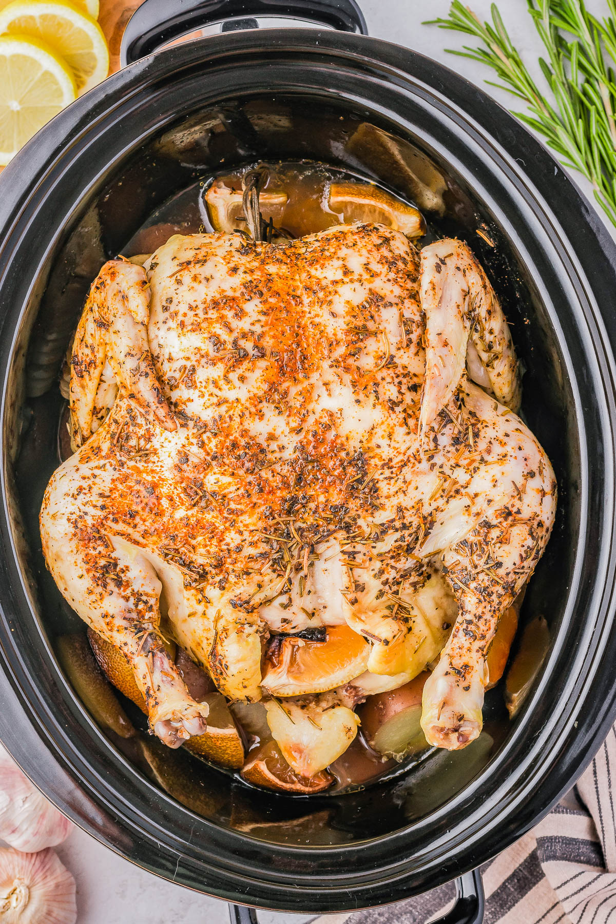 A seasoned whole chicken is cooked in a black slow cooker, surrounded by lemon wedges, garlic cloves, and herbs.