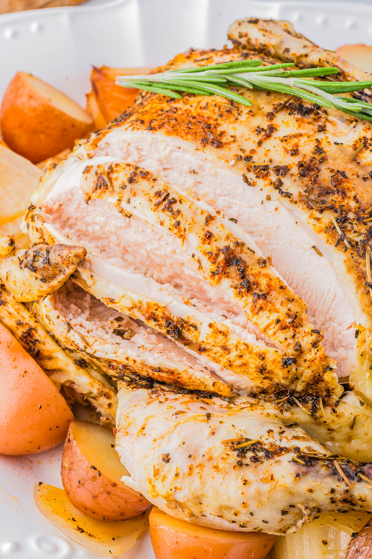 A roasted chicken garnished with rosemary, surrounded by cooked potatoes and onions on a white plate.