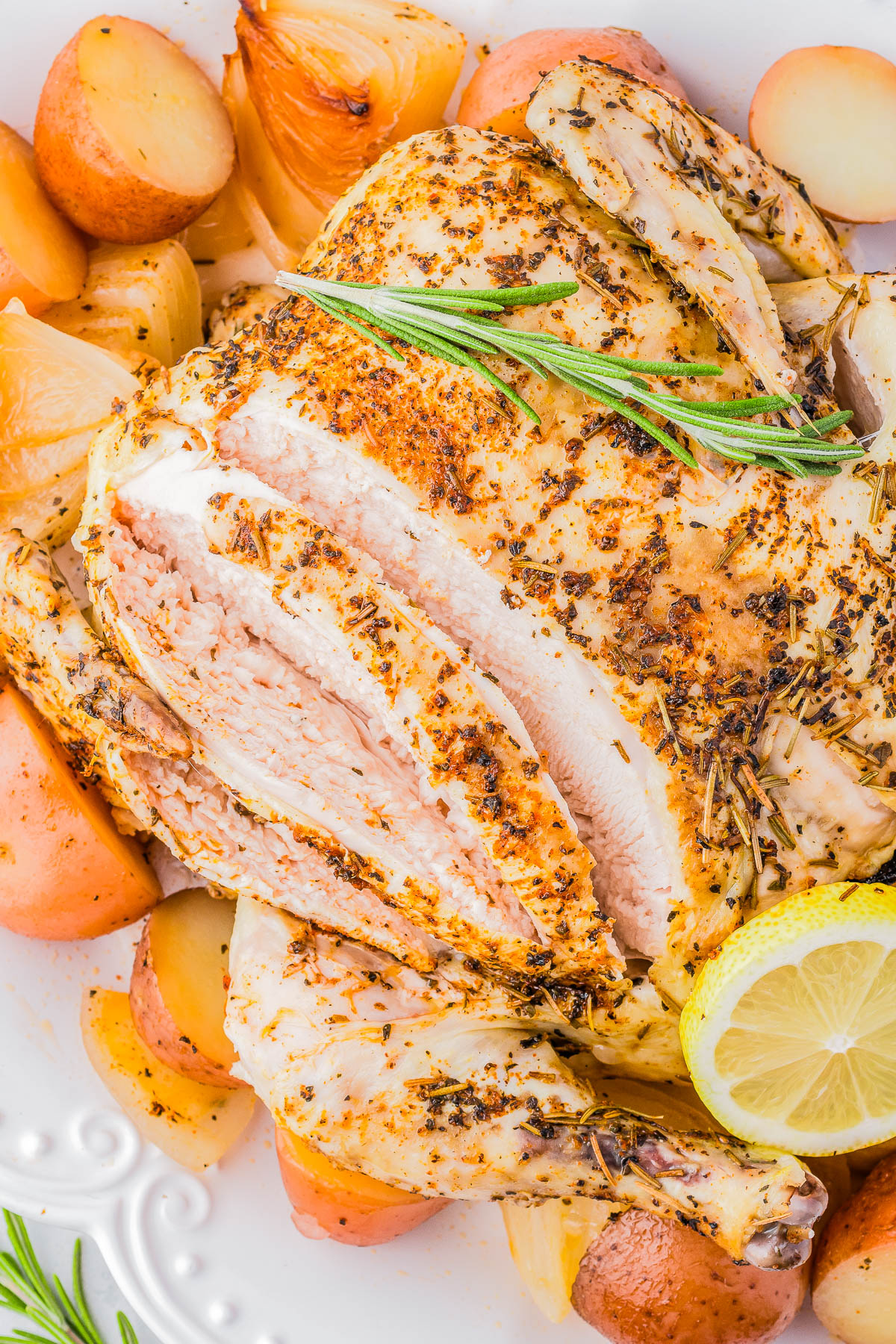 A roasted chicken garnished with rosemary and lemon slices, surrounded by roasted potatoes and onions on a white plate.