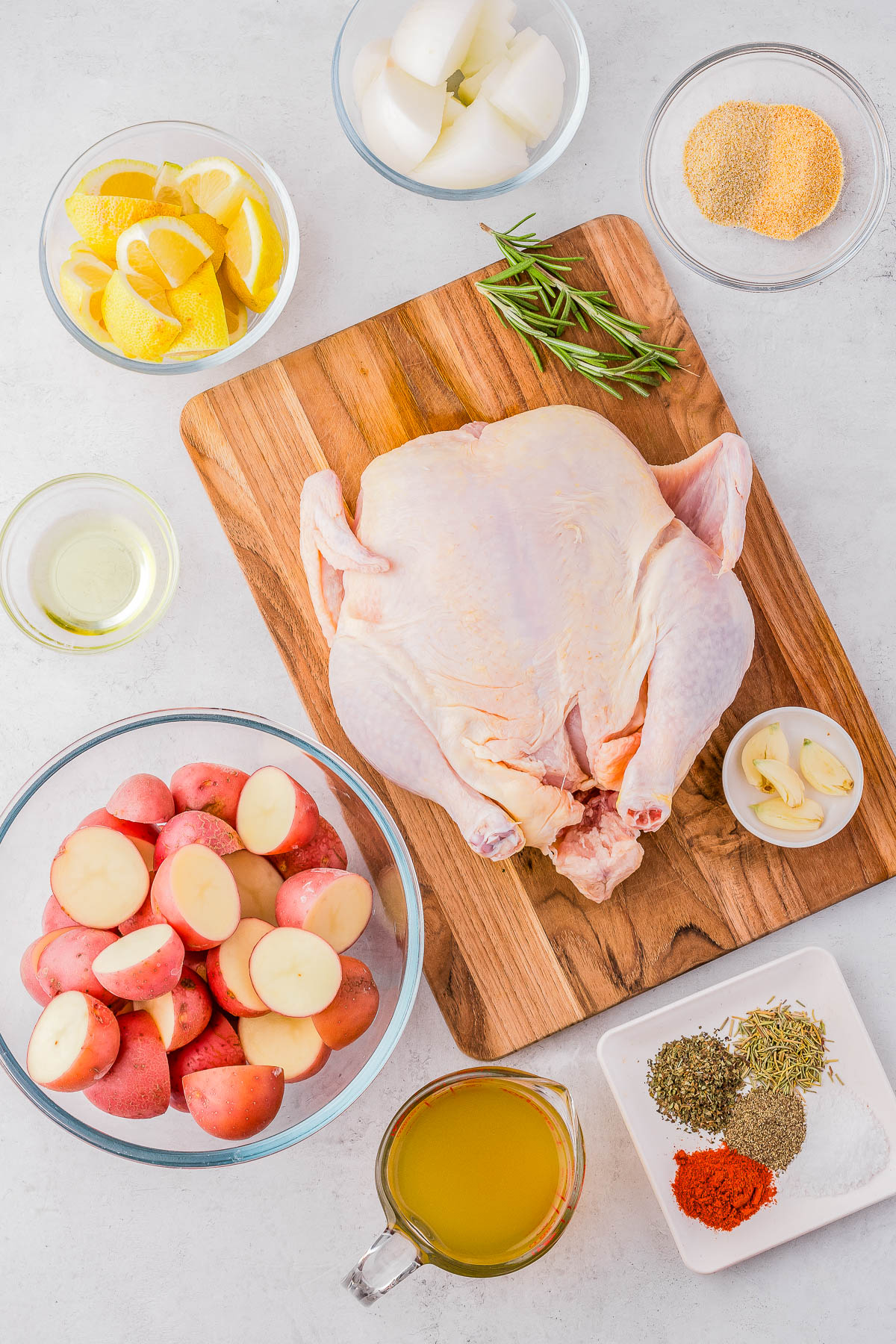 An uncooked whole chicken on a wooden board surrounded by bowls of sliced potatoes, lemon wedges, diced onions, garlic cloves, herbs, spices, olive oil, and chicken broth.