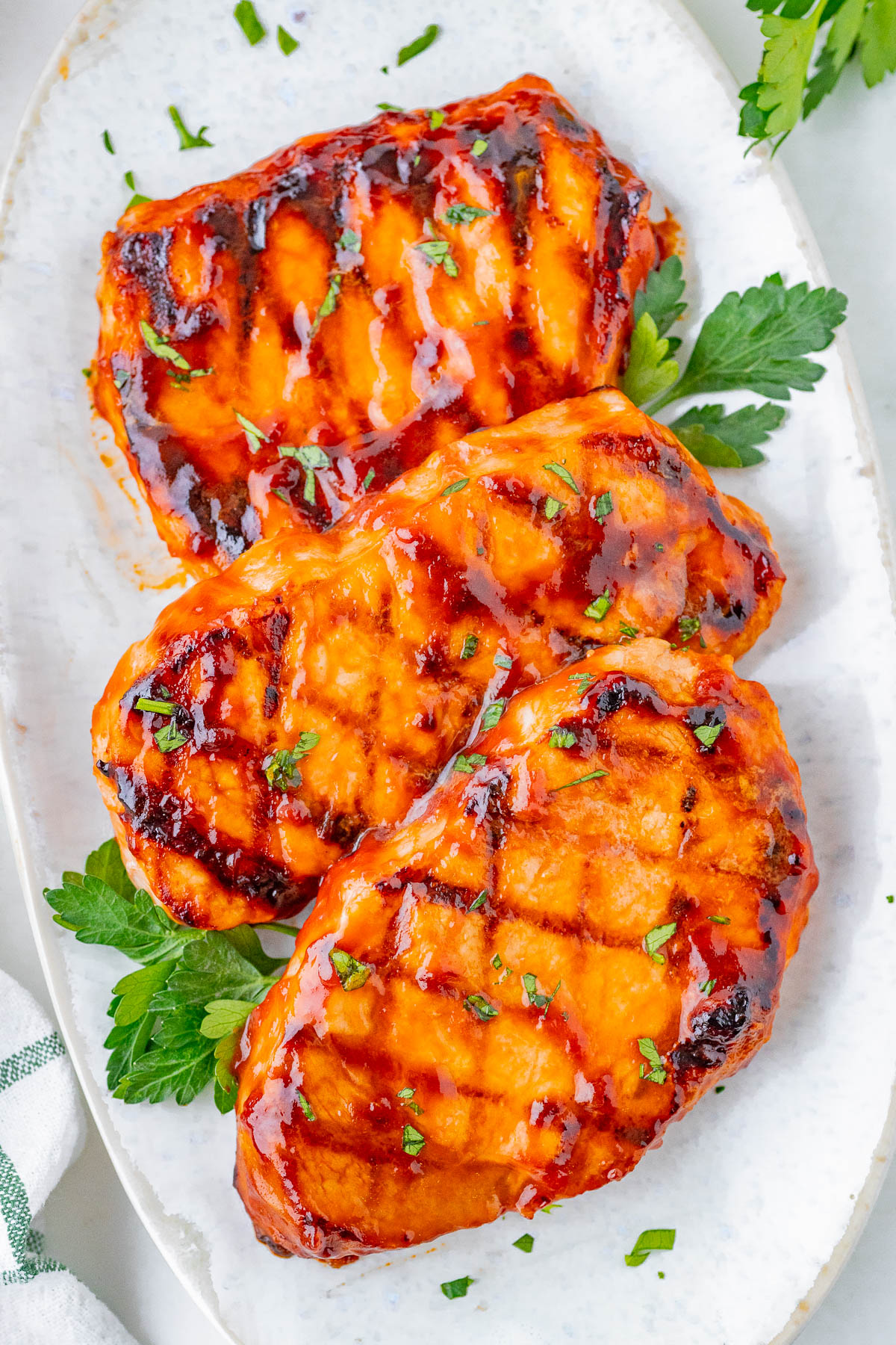 Three grilled, glazed pork chops garnished with chopped parsley, served on a white plate with additional parsley leaves.