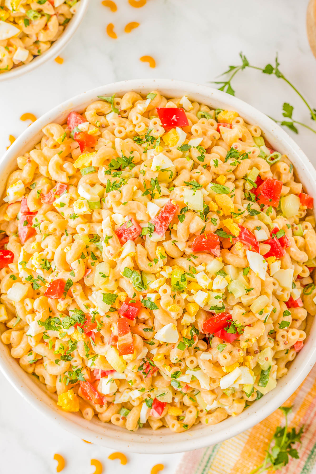 A bowl of macaroni salad with diced tomatoes, yellow bell peppers, chopped eggs, and herbs on a white surface.