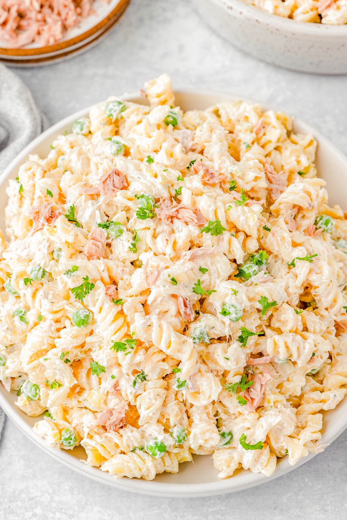 A bowl of creamy tuna pasta salad with rotini pasta, peas, and fresh parsley, placed on a light-colored surface.