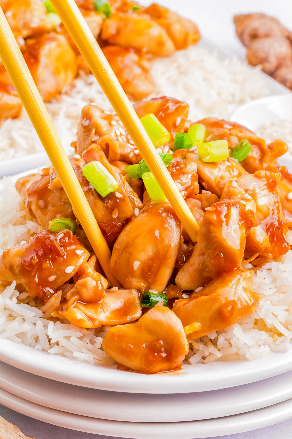 A close-up of a plate of rice topped with saucy chunks of teriyaki chicken, garnished with green onions, with chopsticks picking up a piece of chicken.