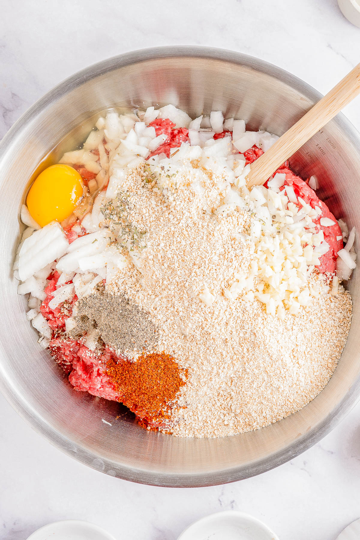 A metal bowl containing raw minced meat, an egg, chopped onions, minced garlic, breadcrumbs, and various spices. A wooden spoon is also in the bowl.