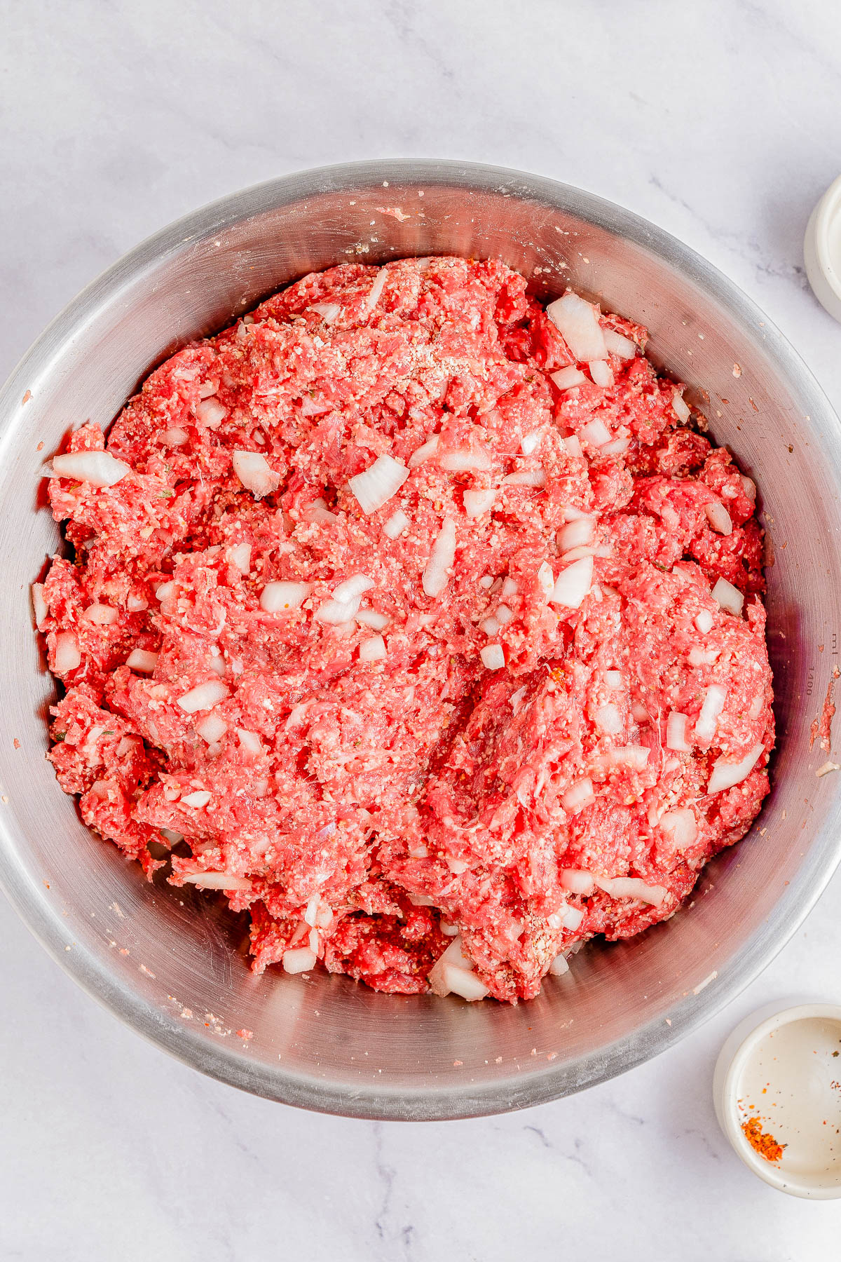 A metal bowl filled with a mixture of ground beef, onions, and spices, seen from above on a light-colored surface.