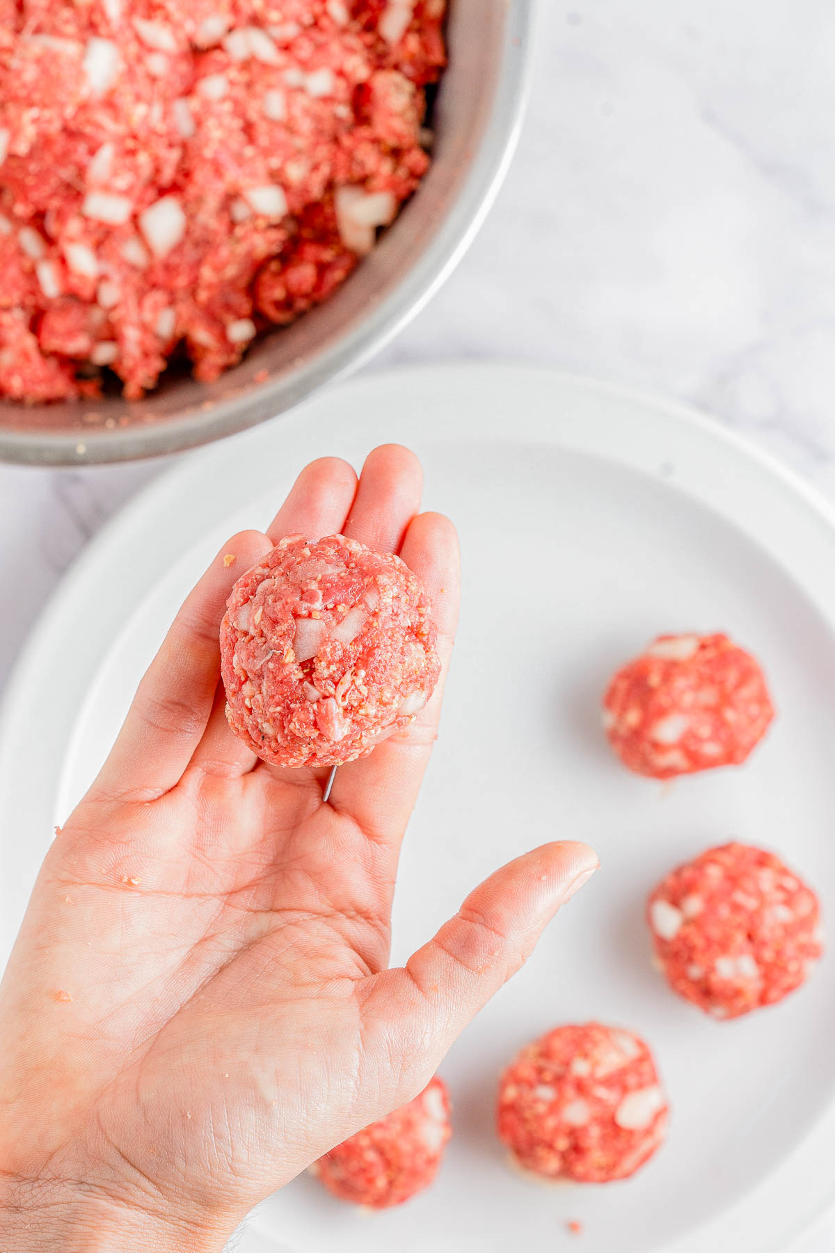 A hand holding a raw meatball over a plate with other uncooked meatballs. A metal bowl with meat mixture is in the background.