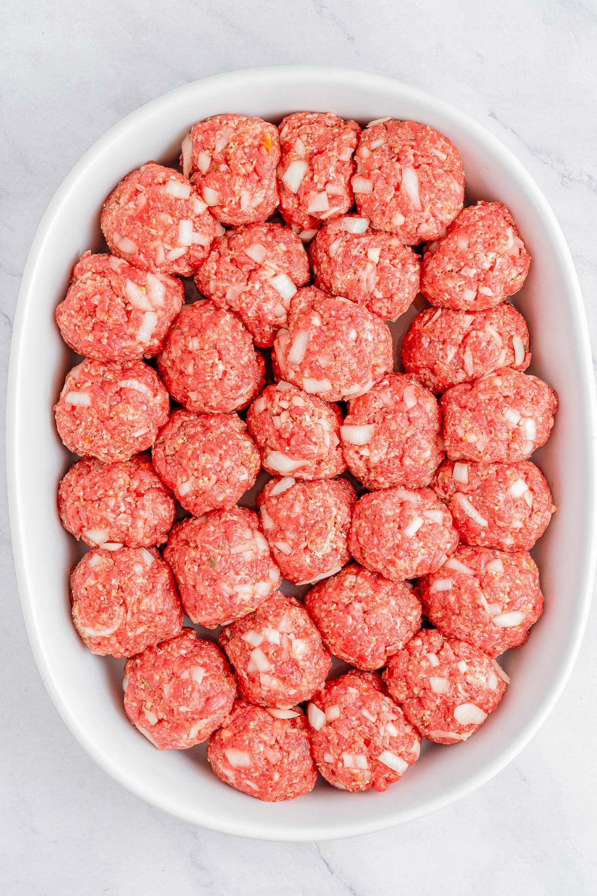 A white dish filled with rows of raw, round meatballs mixed with white onion pieces placed on a light-colored surface.