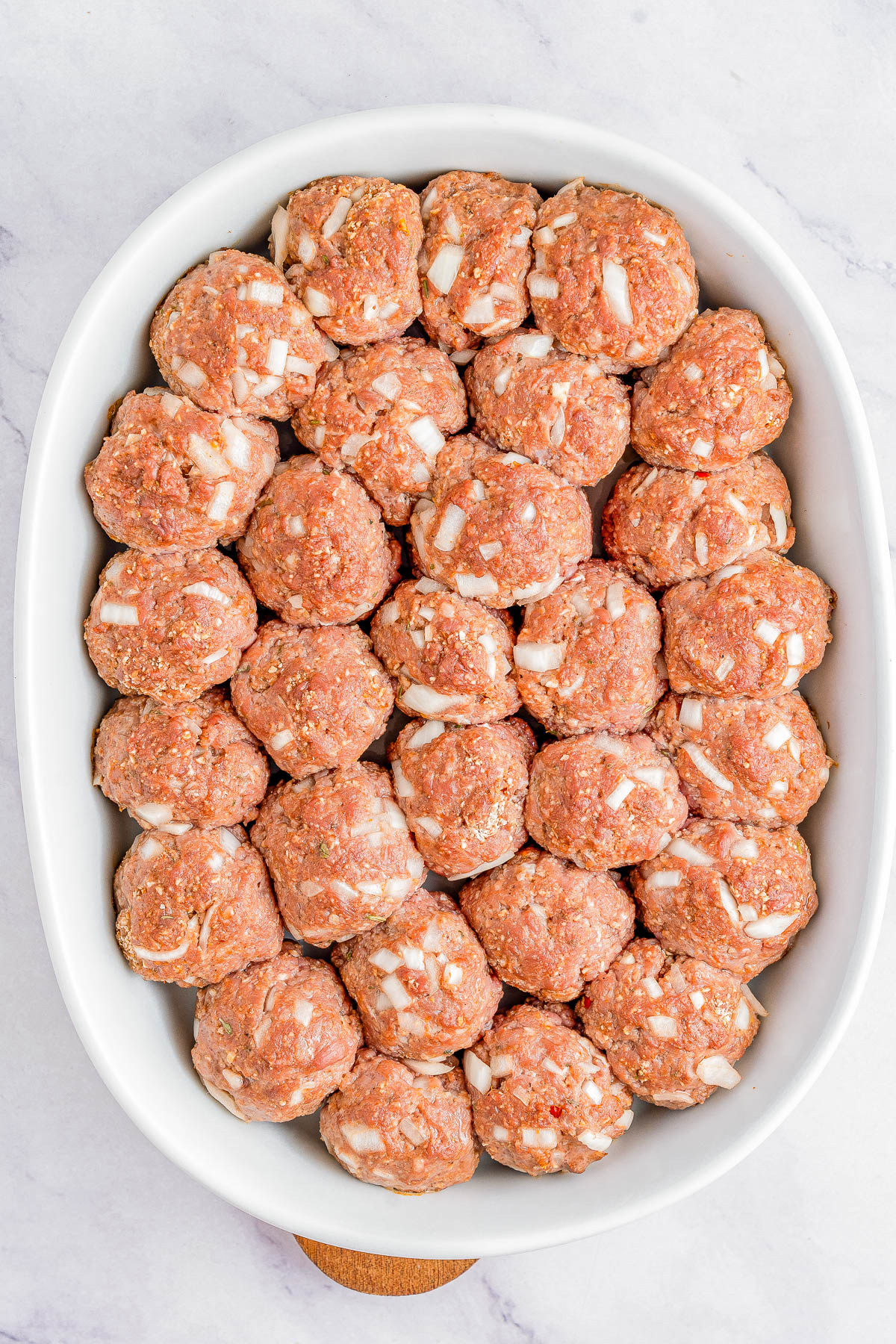 A white dish filled with evenly arranged raw meatballs mixed with finely chopped onions and herbs on a marble surface.