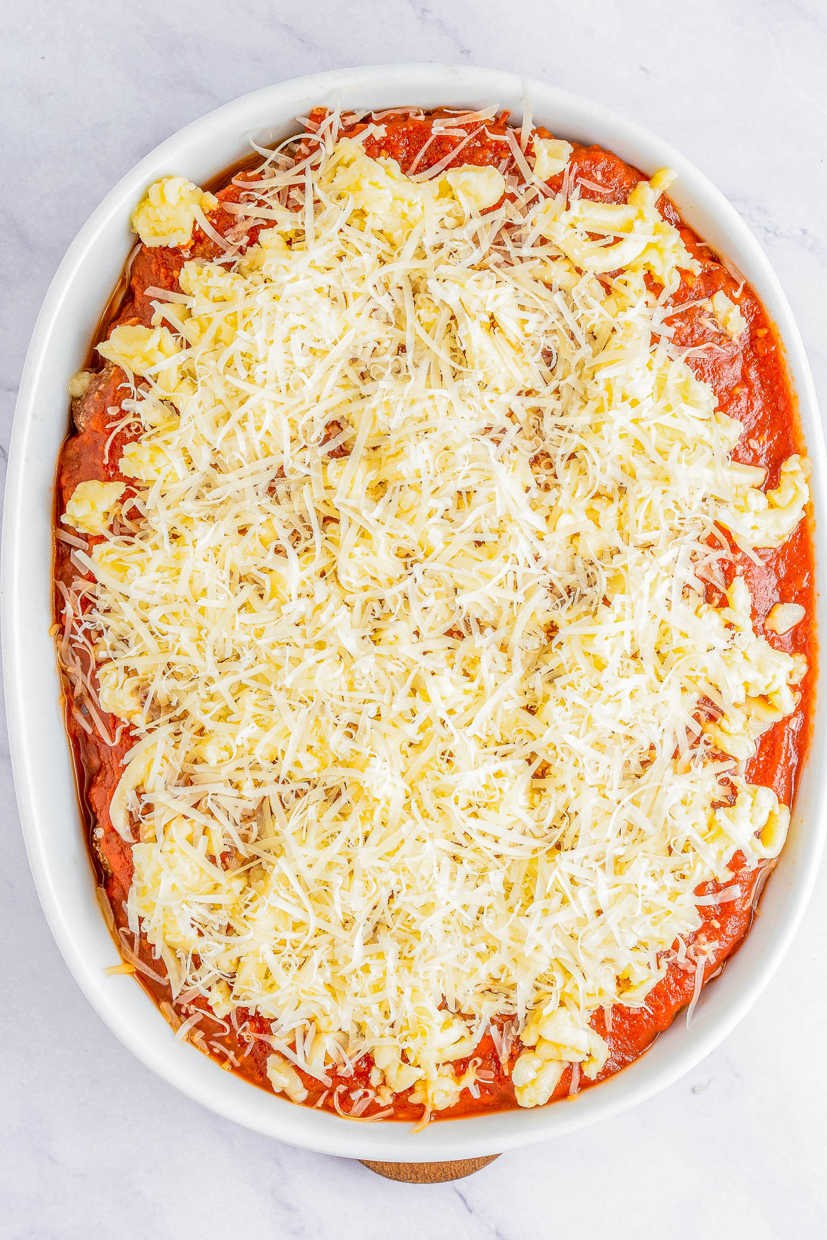 An overhead view of an oval baking dish filled with uncooked lasagna, showing layers of tomato sauce and shredded cheese on top.