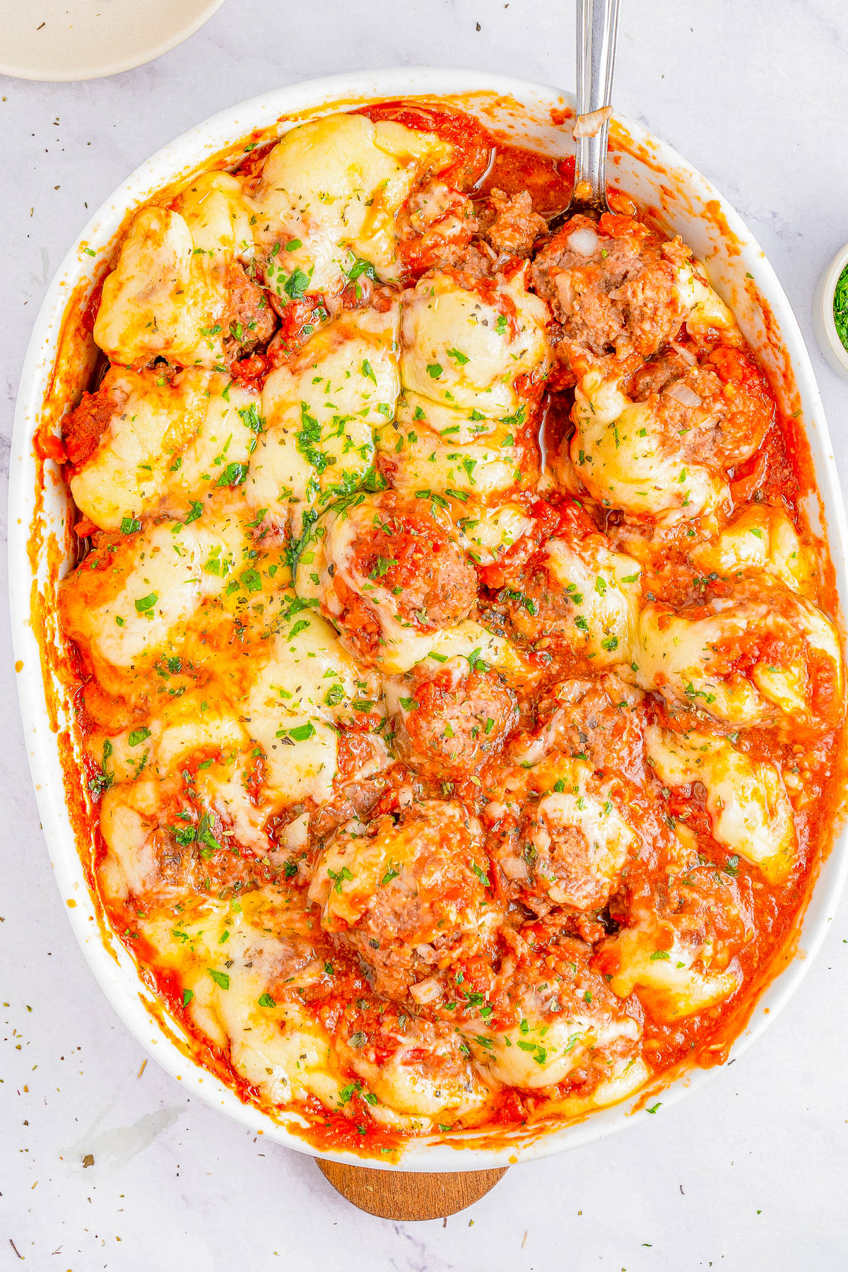 A baked dish of meatballs covered in melted cheese and tomato sauce is garnished with chopped green herbs in a white casserole dish.