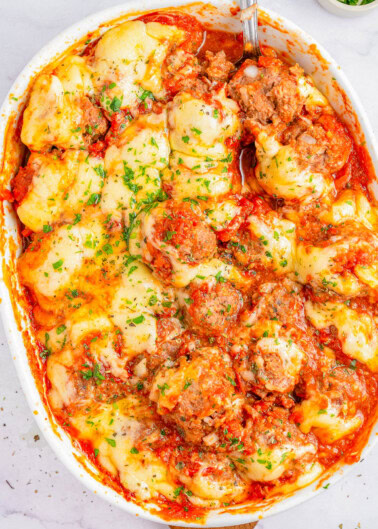 A baked dish in a white casserole pan featuring meatballs, melted cheese, and tomato sauce garnished with chopped parsley. A serving spoon is partially visible on the right side of the dish.