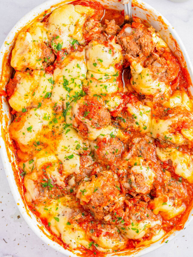 A baked dish in a white casserole pan featuring meatballs, melted cheese, and tomato sauce garnished with chopped parsley. A serving spoon is partially visible on the right side of the dish.