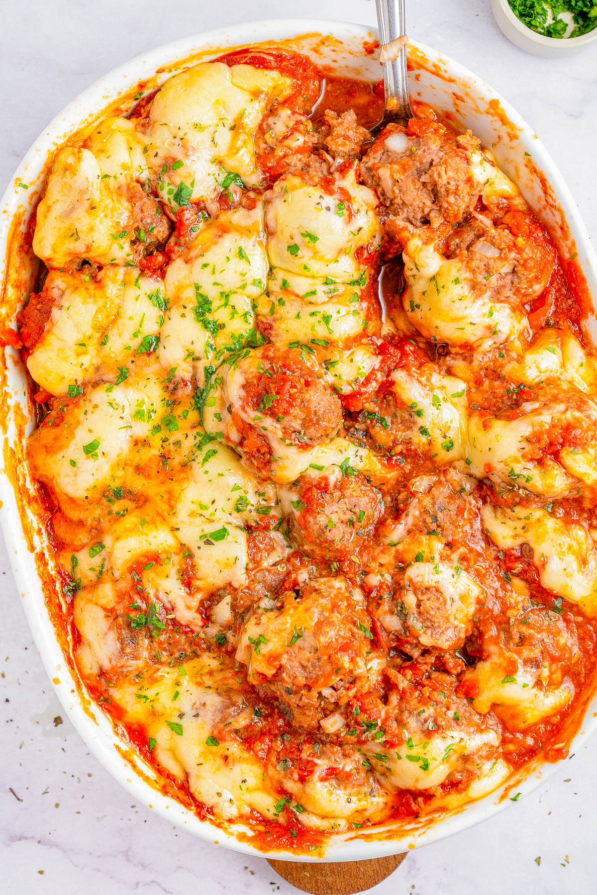 A baked dish in a white casserole pan featuring meatballs, melted cheese, and tomato sauce garnished with chopped parsley. A serving spoon is partially visible on the right side of the dish.