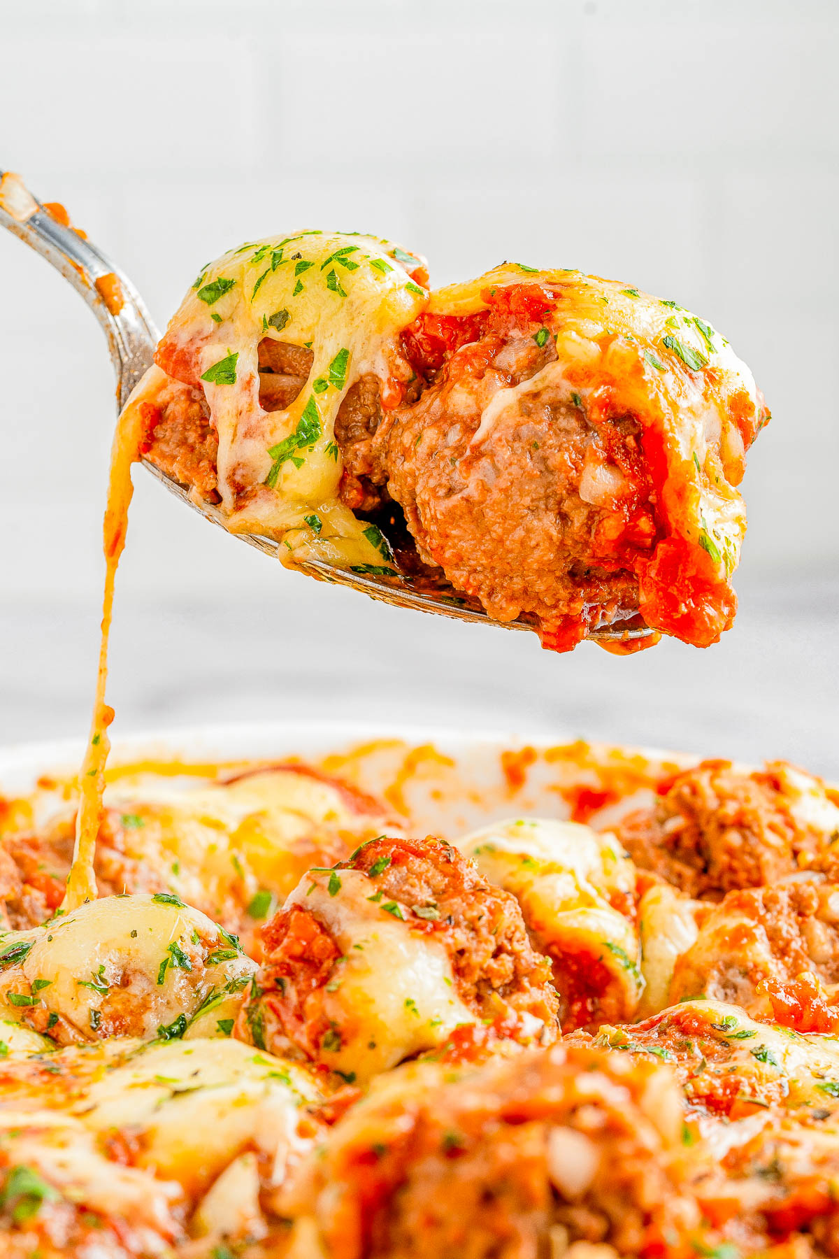 Close-up of a spoon lifting a cheesy meatball from a dish filled with meatballs, melted cheese, and tomato sauce, garnished with chopped herbs.