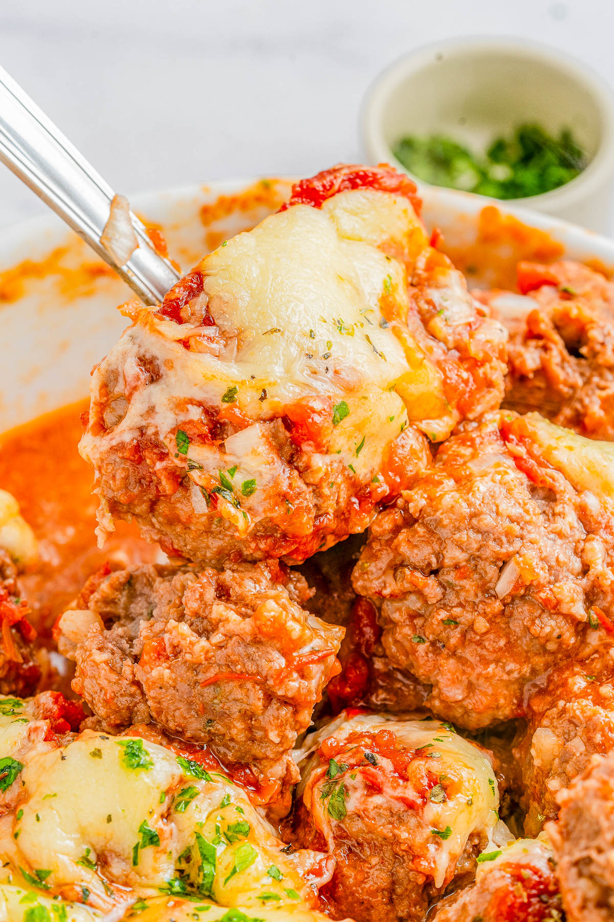 A close-up of melted cheese-covered meatballs in tomato sauce, garnished with chopped parsley.