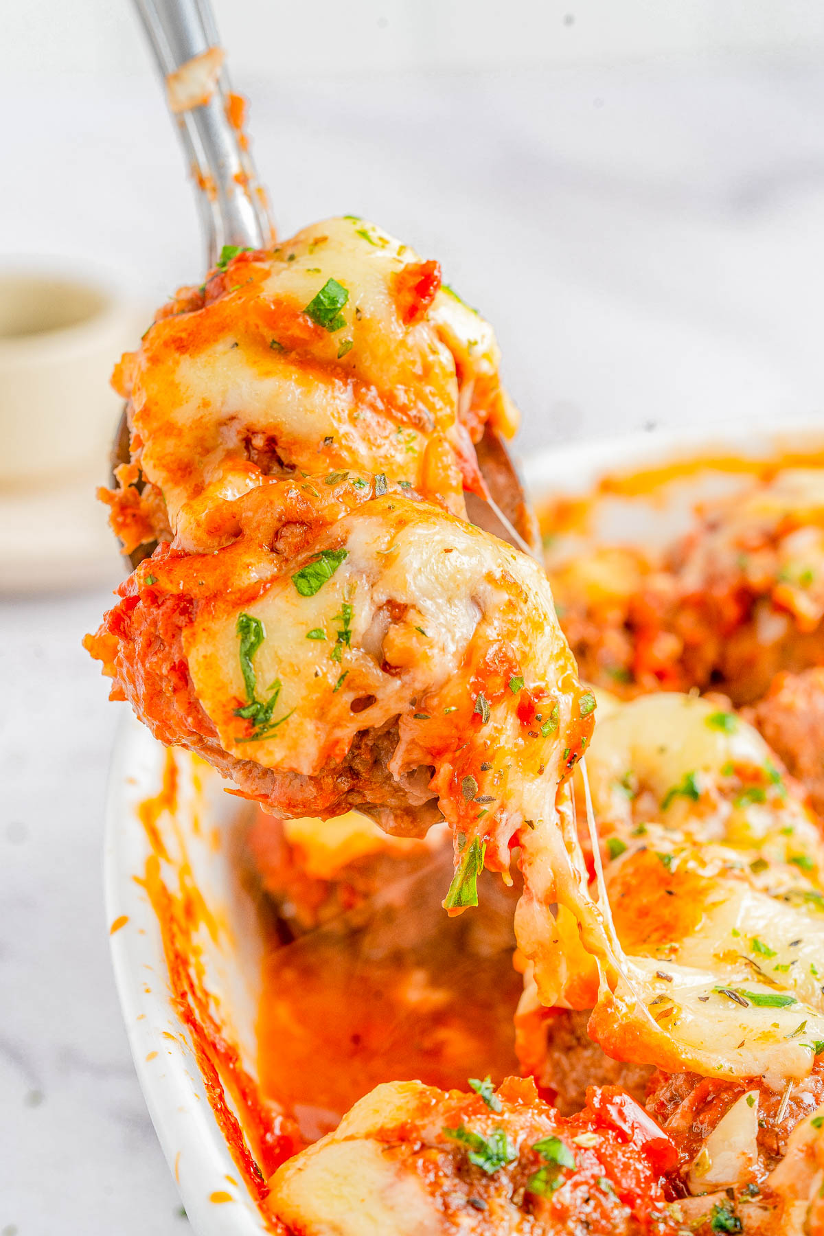 A spoon lifts a portion of cheesy, saucy dish with herbs from a white baking dish, showcasing melty cheese and chunky ingredients.