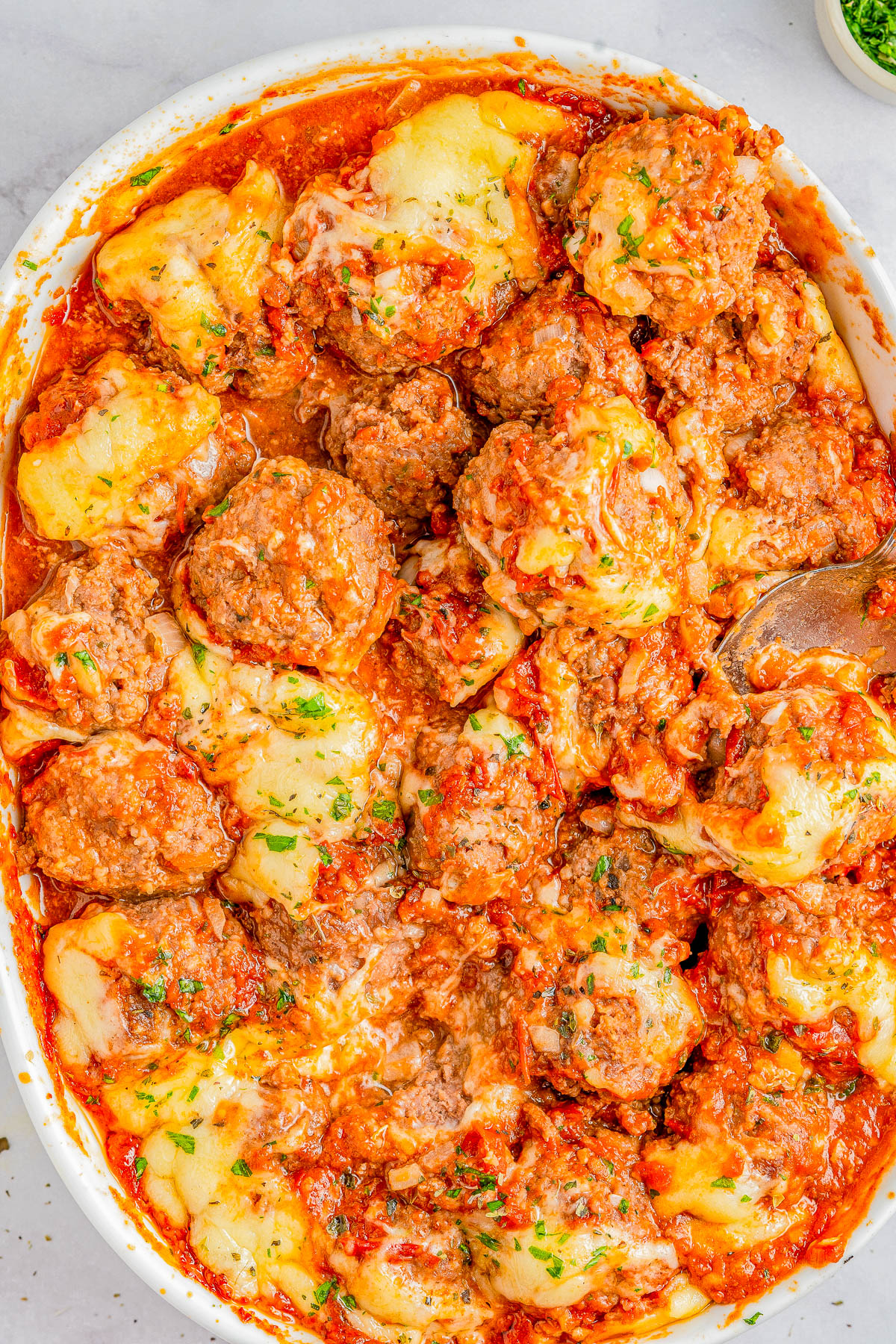 A baked dish with meatballs, cheese, marinara sauce, and herbs in a white oval dish.