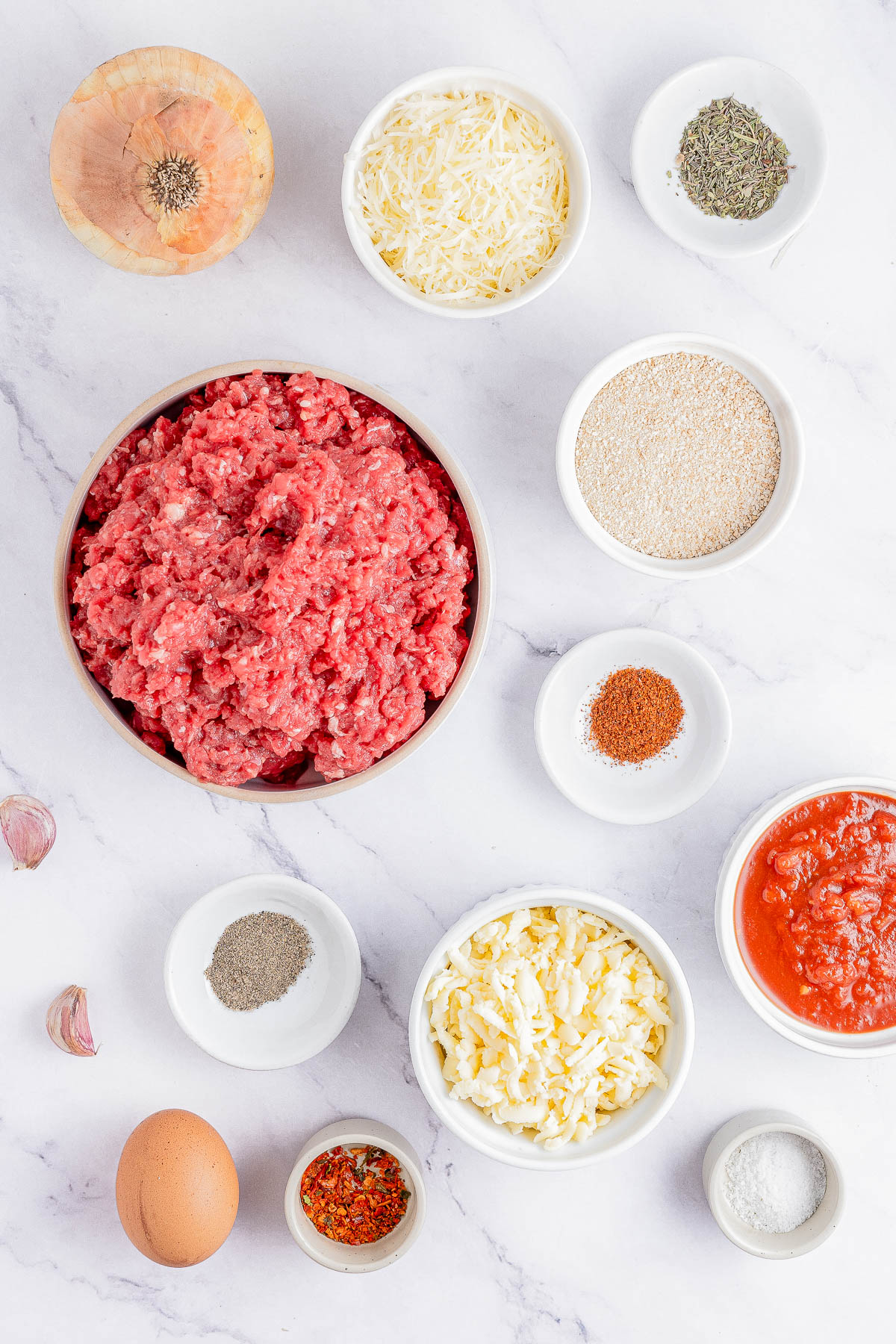 Image showing various ingredients for a meatball recipe including ground meat, onions, garlic, egg, spices, cheese, breadcrumbs, and tomato sauce, all arranged in small bowls on a marble surface.