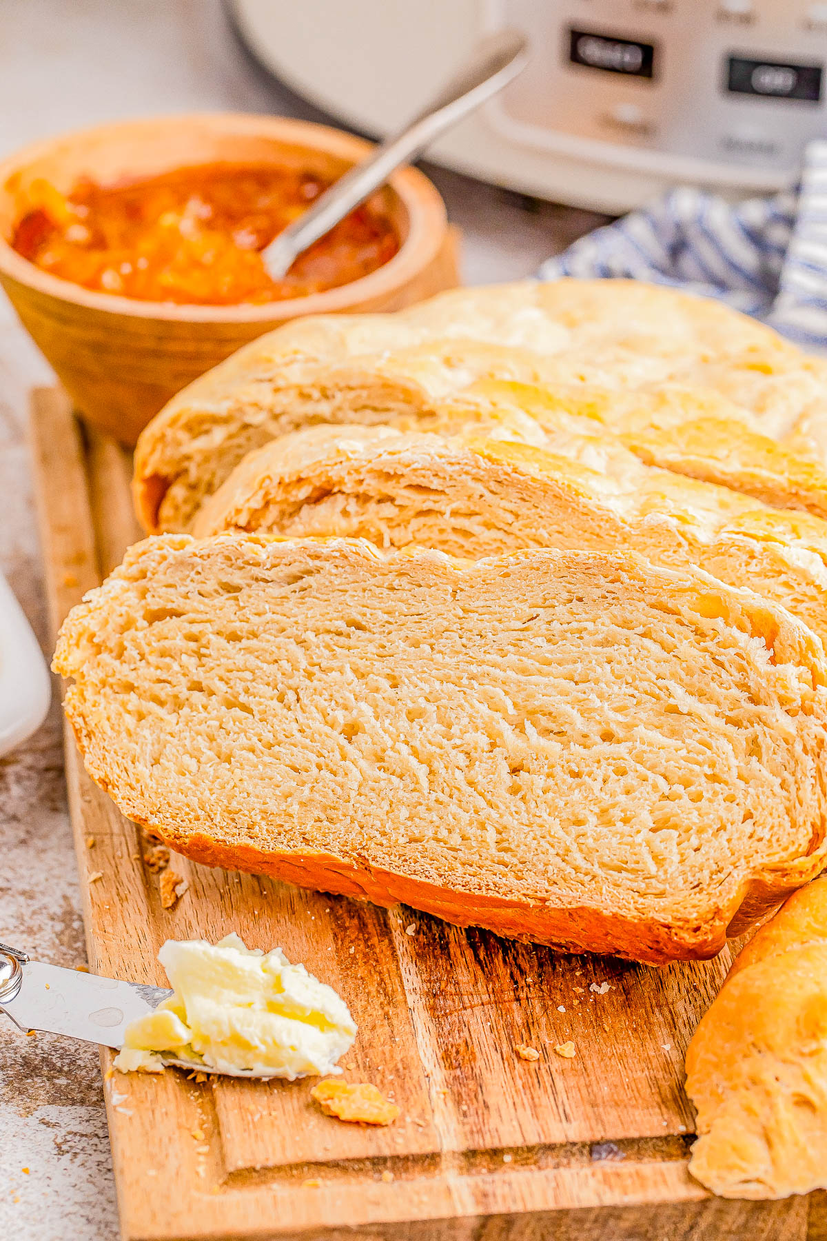 A sliced loaf of bread on a wooden cutting board with a knife spread with butter. A bowl of jam with a spoon is in the background.