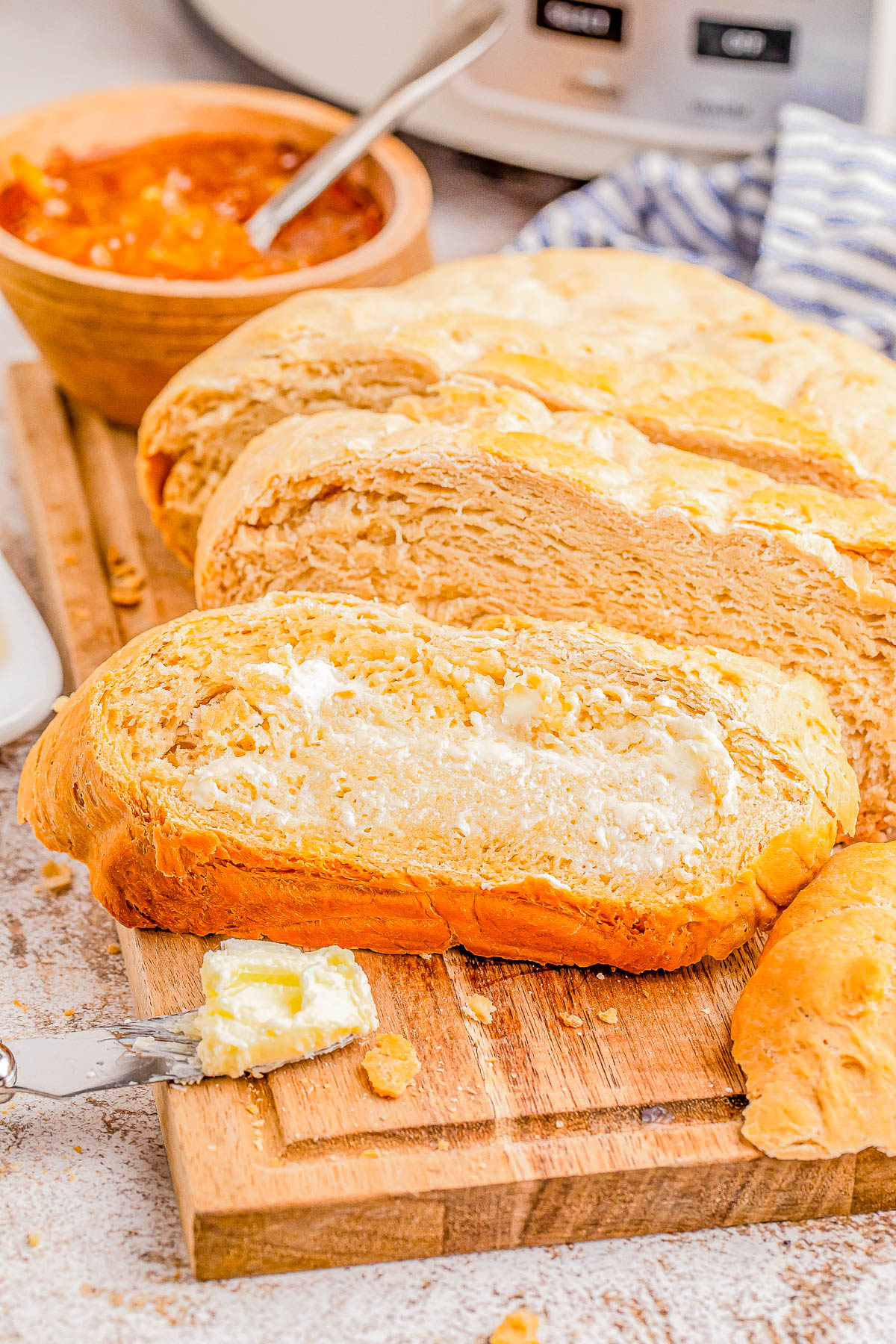 Freshly sliced bread with butter on a wooden board, accompanied by a knife and a bowl of orange marmalade in the background.