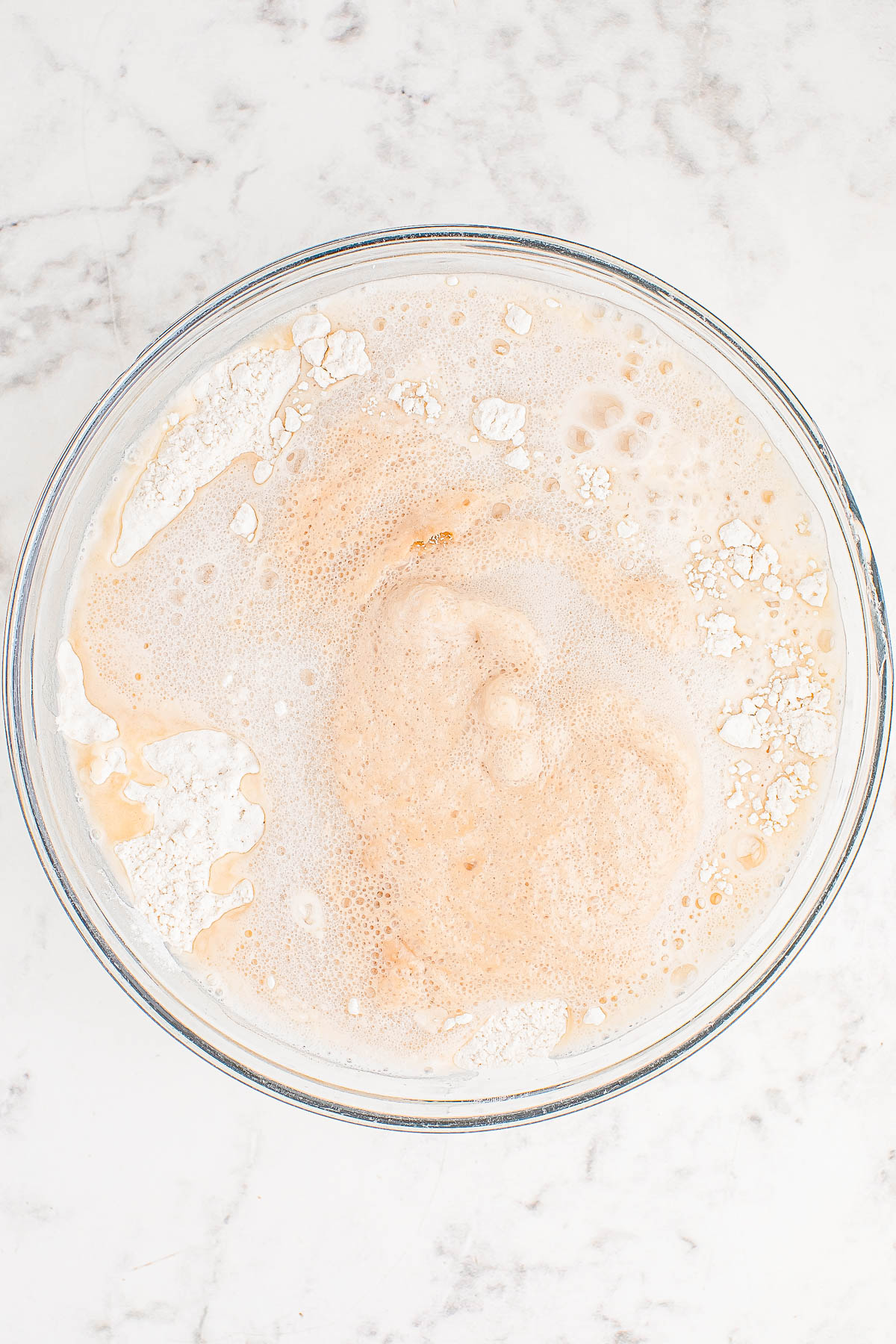 A glass bowl contains a frothy mixture with visible pockets of liquid and patches of dry powder on a light marble surface.