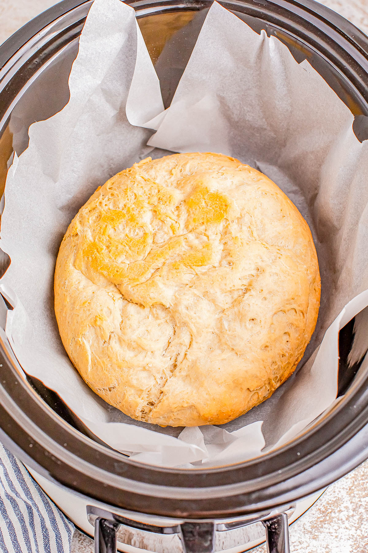 A round loaf of bread rests on parchment paper inside a slow cooker. The exterior is golden brown and appears freshly baked.