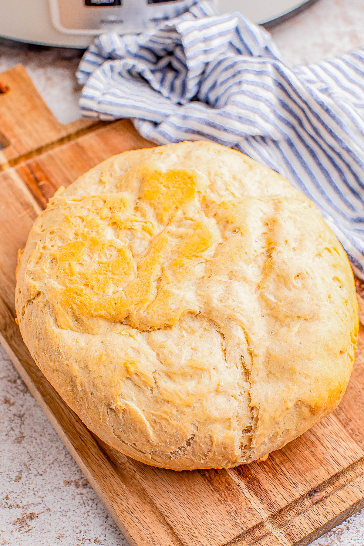 A round loaf of freshly baked bread sits on a wooden cutting board, with a striped cloth in the background.