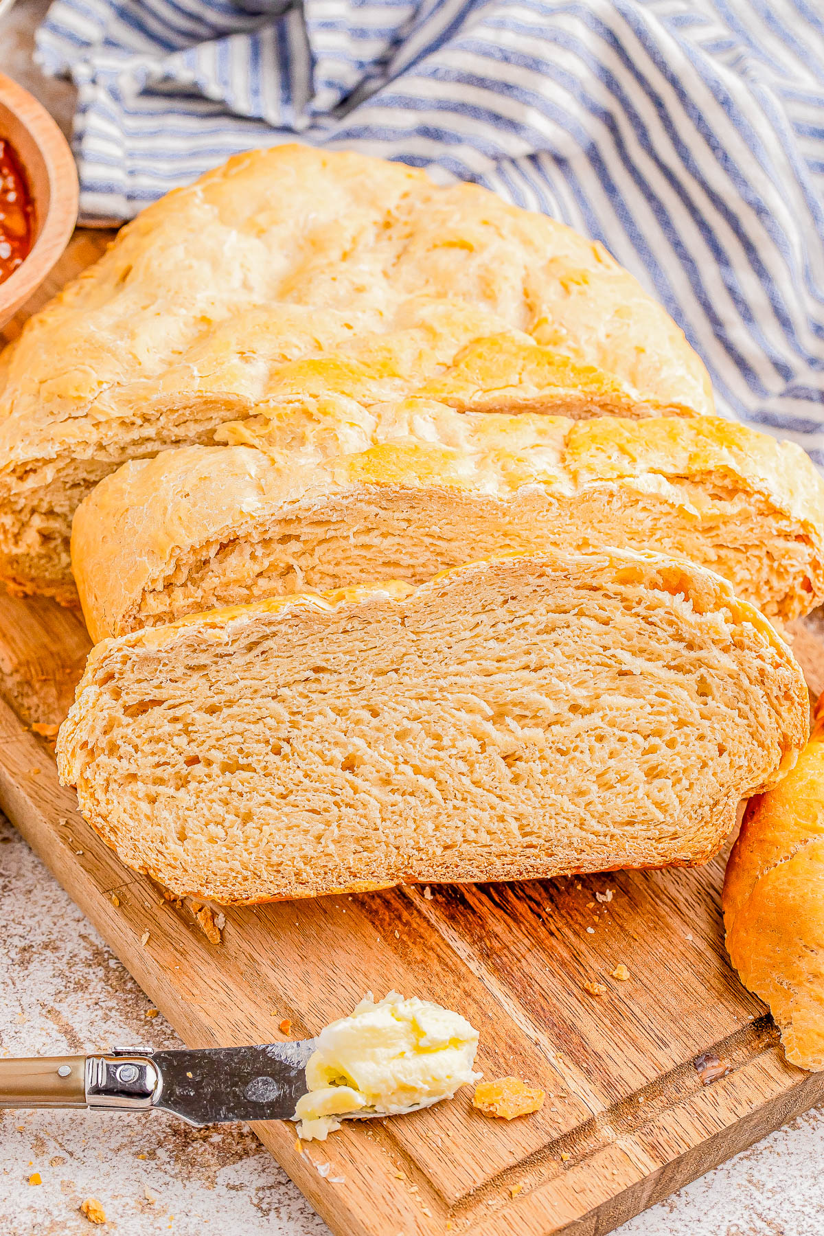 A sliced loaf of bread on a wooden cutting board with a knife spreading butter. A striped cloth and a bowl of sauce are in the background.