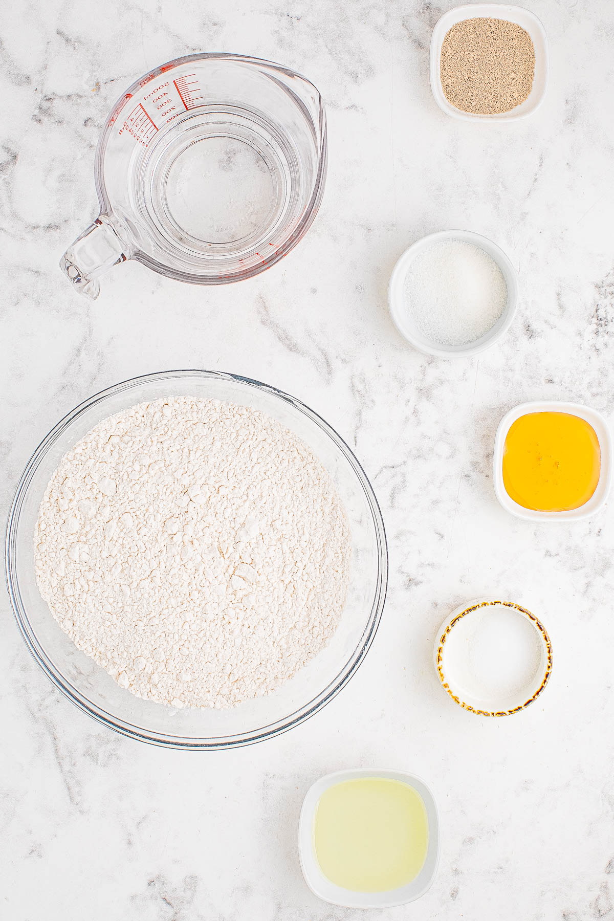 Top-down view of baking ingredients on a marble surface, featuring flour, water, yeast, sugar, salt, oil, and an egg yolk in separate containers.