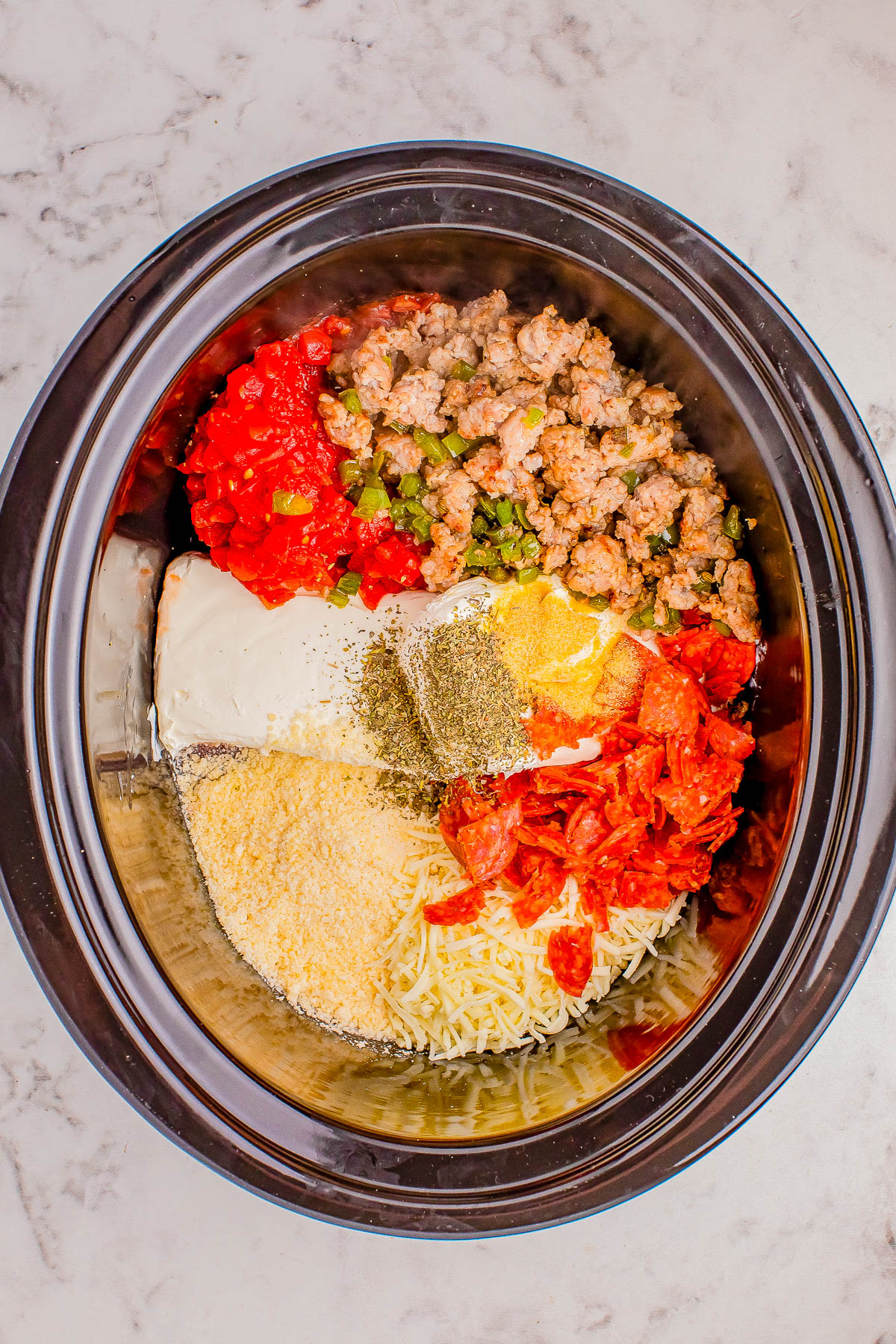 A crockpot filled with diced tomatoes, cooked sausage, chopped green bell peppers, shredded cheese, pepperoni slices, cream cheese, and various seasonings on a marble countertop.