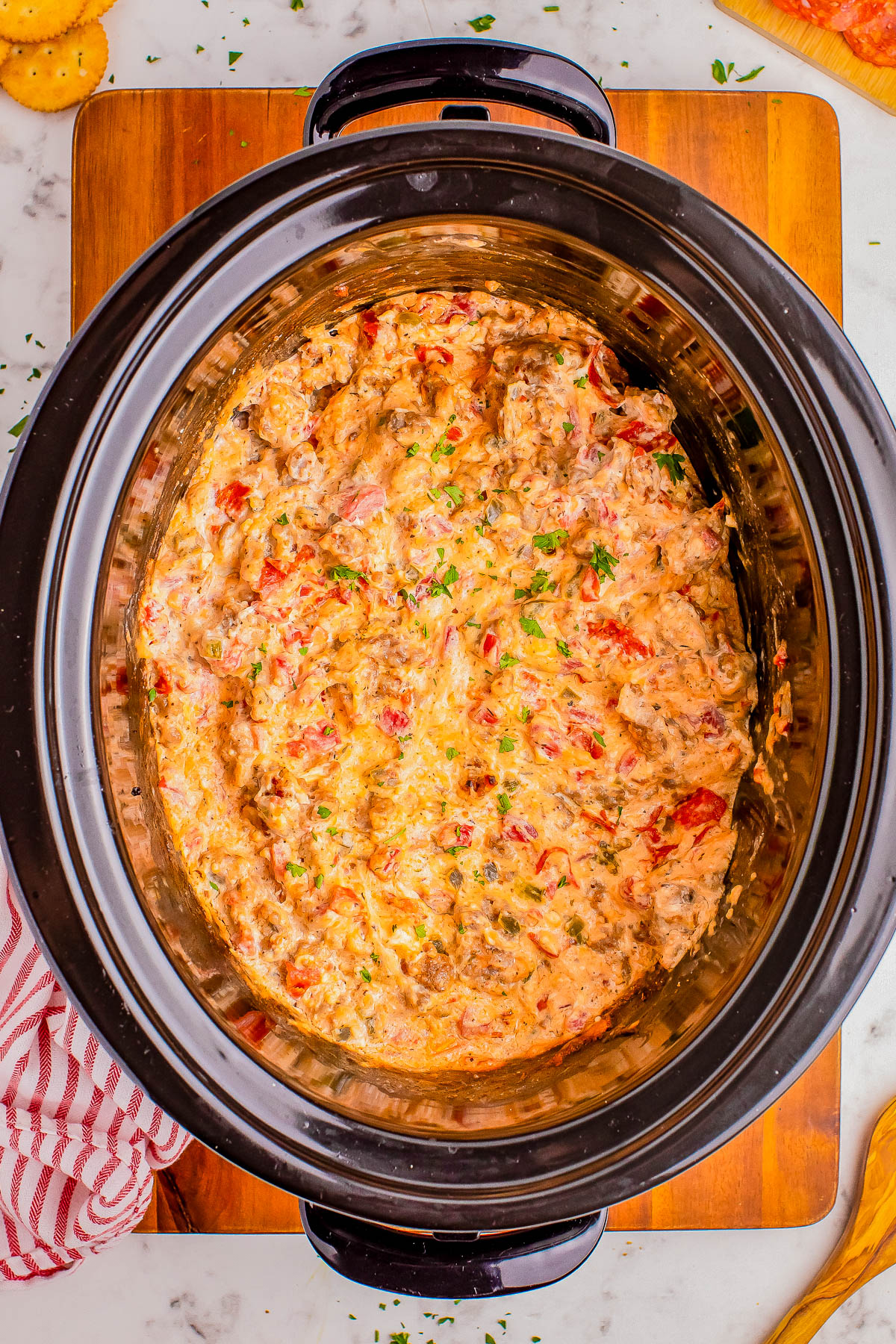 A slow cooker filled with a creamy, mixed dish containing chunks of meat, tomatoes, and garnished with herbs placed on a wooden surface.